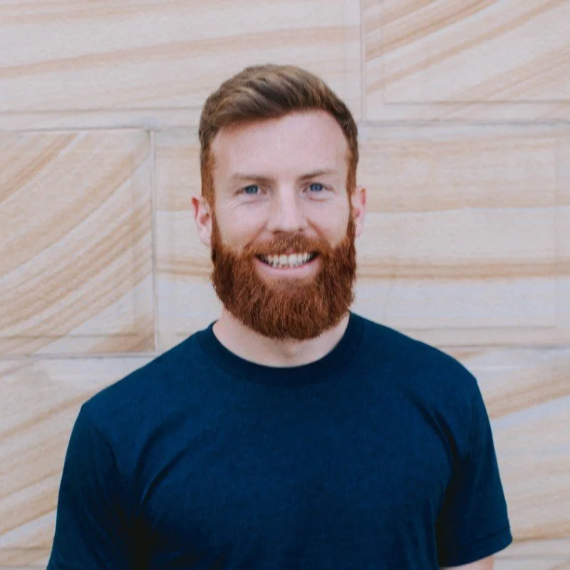 A man with red hair and a full beard smiling, wearing a dark blue t-shirt, standing in front of a beige, wooden textured wall.
