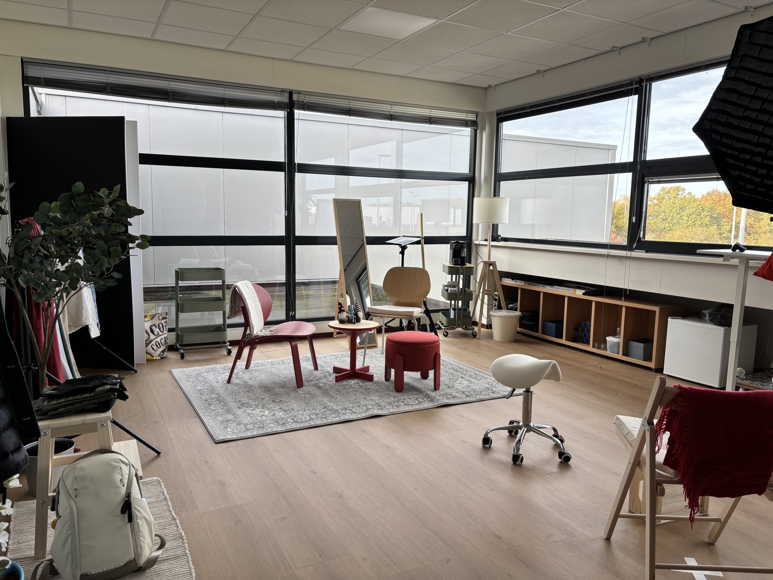 Mijn fotostudio met grote ramen die uitzicht bieden op de buitenwereld. In het midden bevinden zich twee rode stoelen, een kleine tafel met decoraties en een kleine rode kruk.