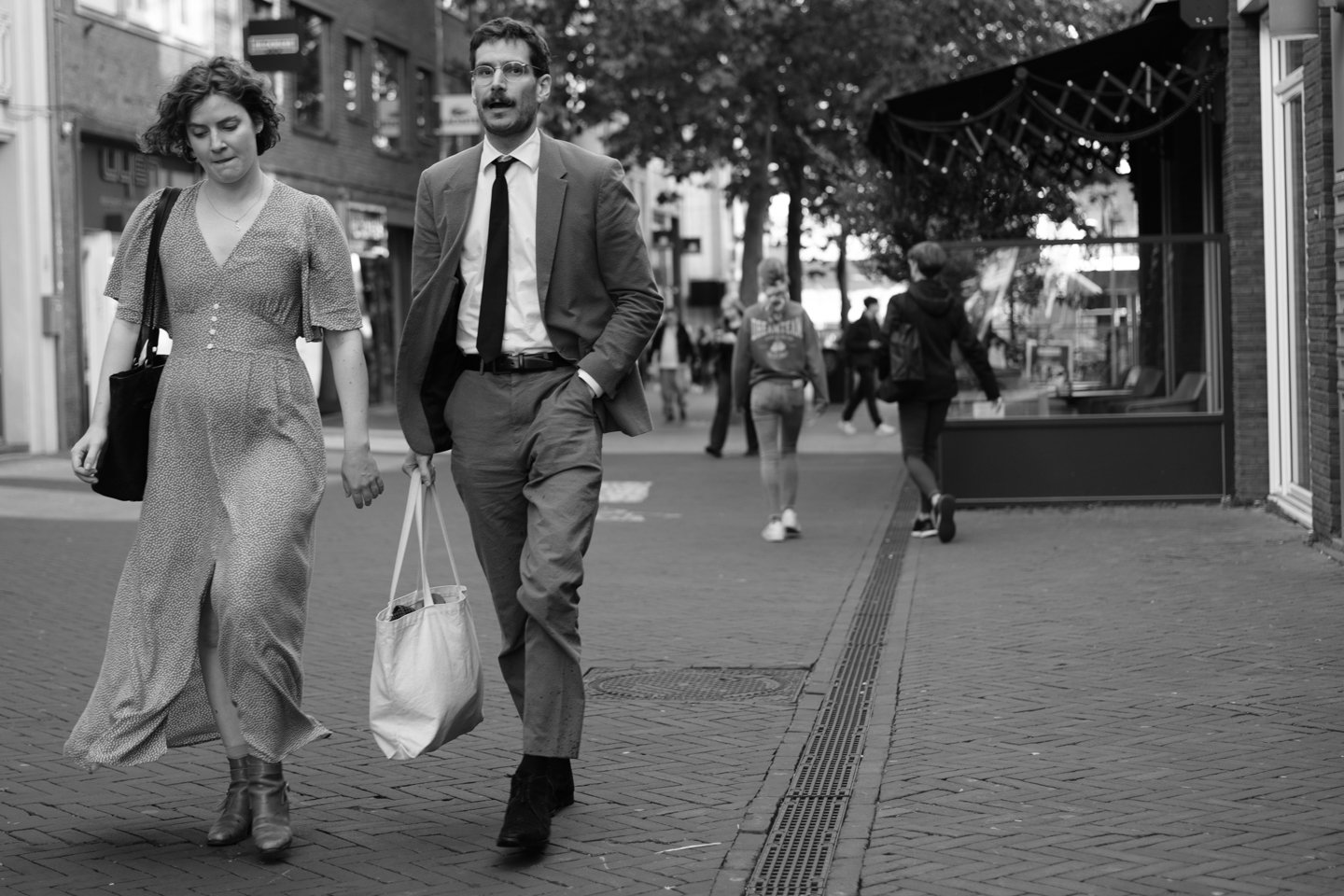 Twee mensen lopen samen op een stadsstraat, de vrouw draagt een lange jurk en laarzen, de man draagt een pak en houdt een tas.