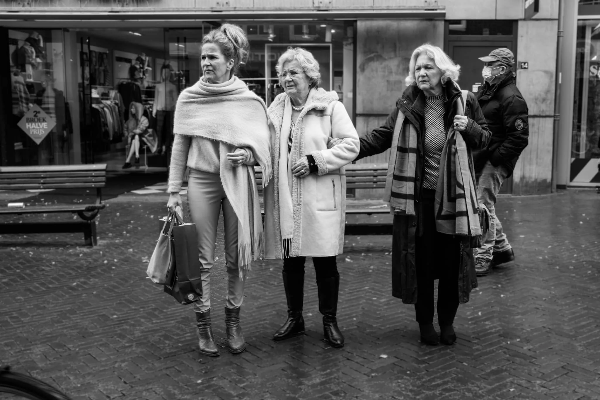 Drie oudere vrouwen staan buiten op een straat, de ene vrouw ondersteunt de andere. Ze dragen allemaal warme jas en schoenen, en één vrouw draagt een gezichtsmasker. Een man met een gezichtsmasker staat op de achtergrond.