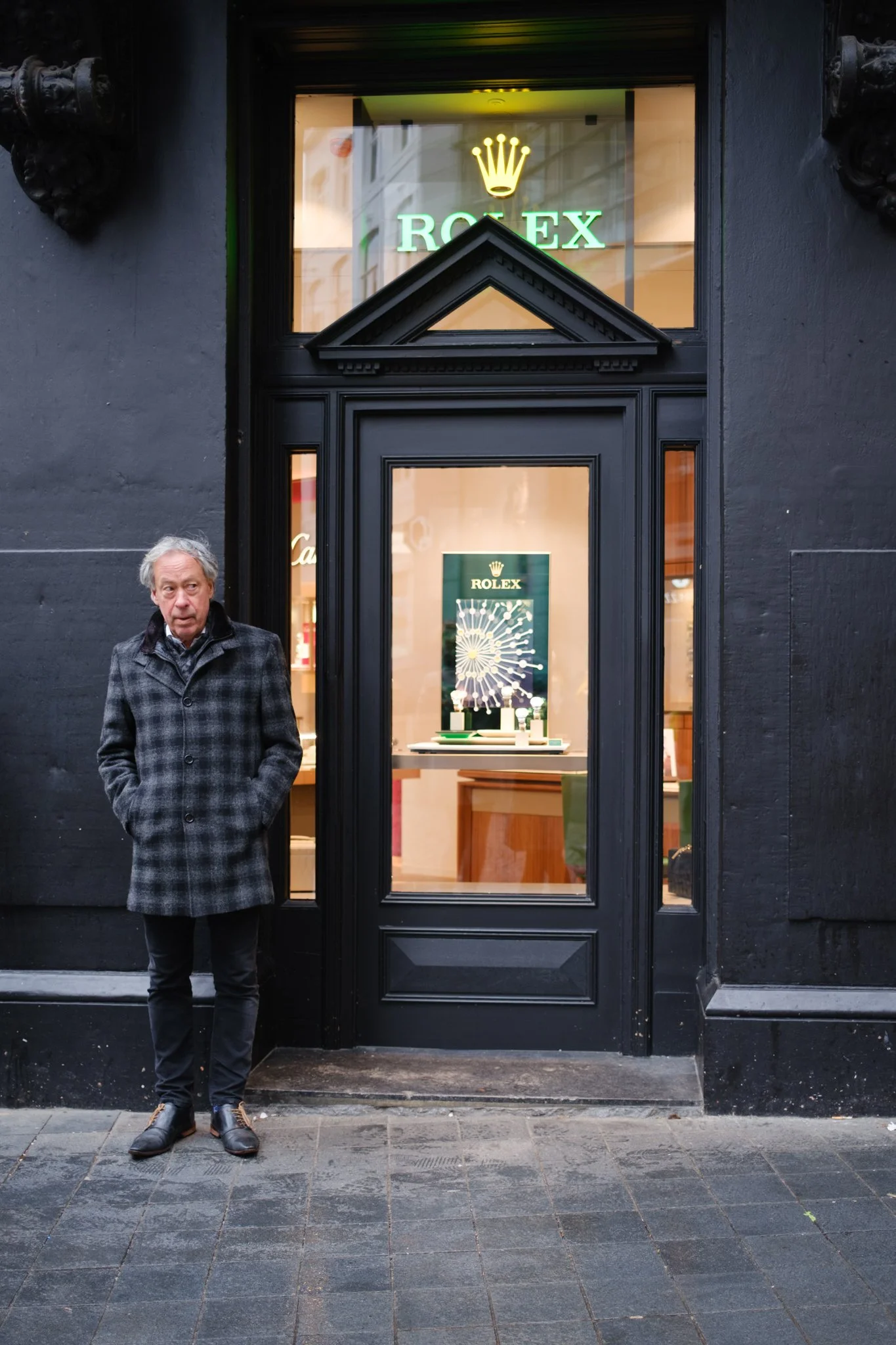 Een man staat buiten een Rolex winkel met een zwarte gevel en grote ramen, binnenin is een Rolex reclame met een horloge te zien.