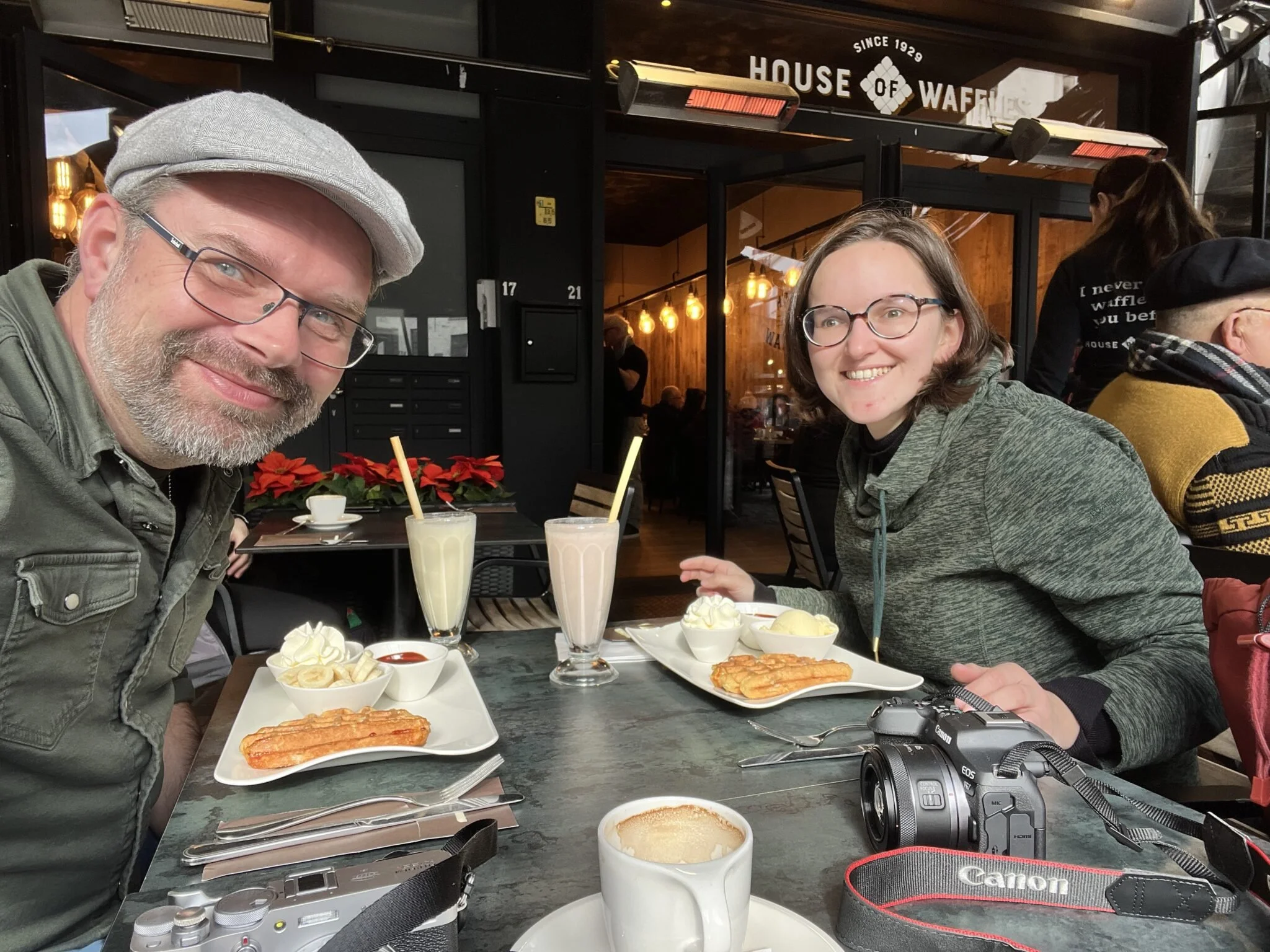 Twee mensen zitten in een restaurant, eten en drinken, met een camera en een fotocamera op tafel.