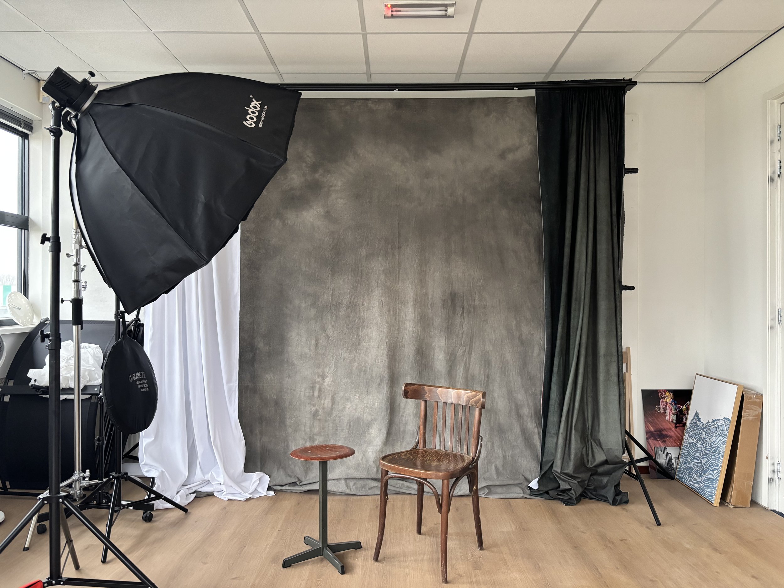Mijn fotostudio met een grijze achtergrond, een houten stoel, een kleine ronde kruk en enkele foto's op de vloer in de hoek. Flitser en een softboxverlichting op een statief te zien.