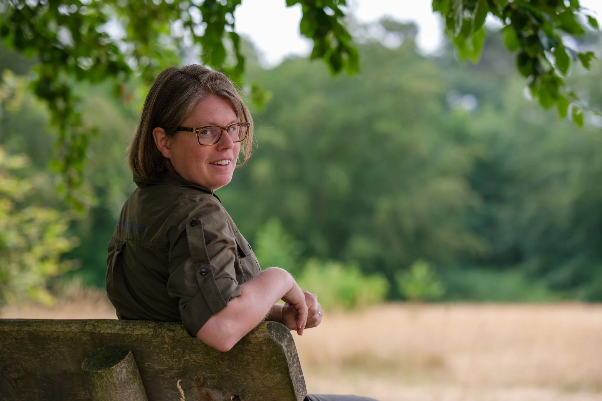 Vrouw met bruin haar en bril zit op een houten bankje in een groen park, kijkt richting camera met een glimlach.