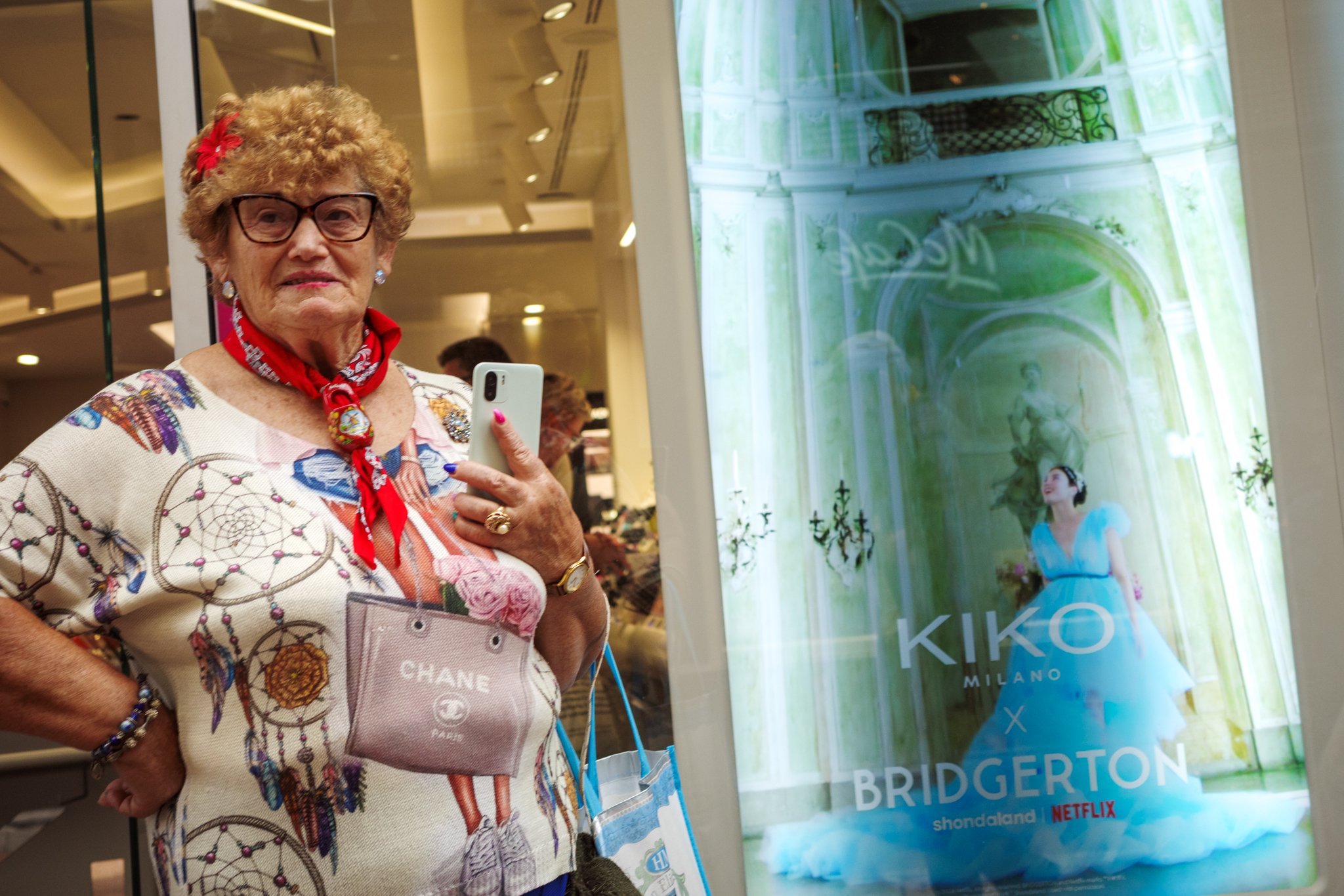 Een oudere vrouw met gekruld haar, brillen, rood sjaaltje, een fauteuil met een merk label CHANEL, die een smartphone vasthoudt, naast een digitale poster van een Netflix serie 'Bridgerton'.