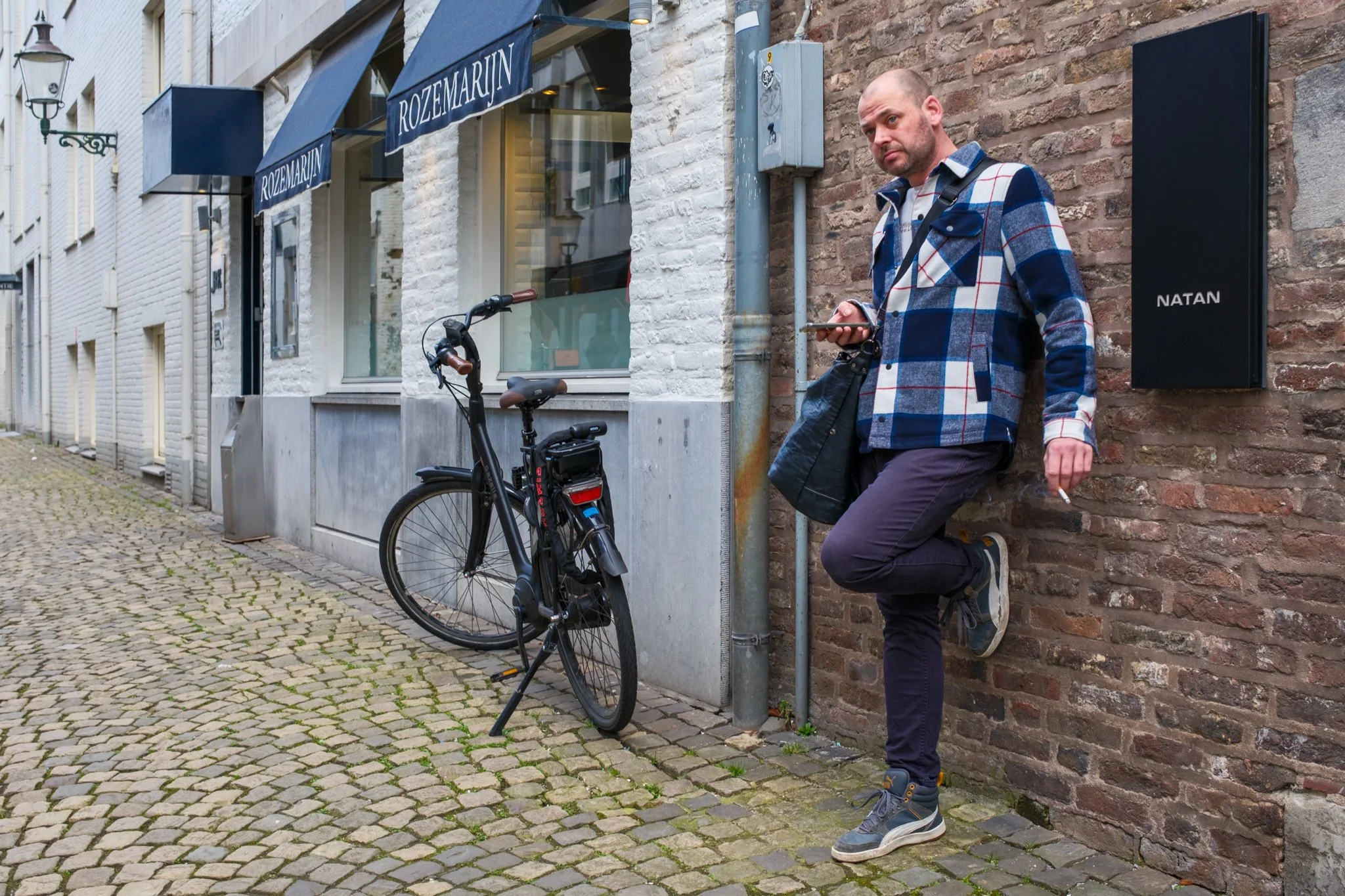 Man leunt tegen een bakstenen muur, houdt een smartphone in zijn hand en heeft een fiets naast zich. Hij draagt een blauwe en grijze geruite jas, donkere pants, en sneakers, op een kasseienstraat voor een winkel met blauwe awning en witgeschilderde g