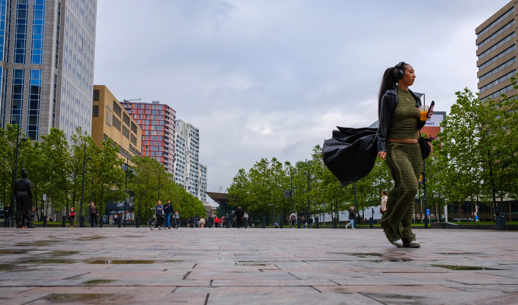 Een vrouw met lange zwarte haar draagt headphones, een groene outfit en een zwart jasje, loopt door een stedelijk plein met gebouwen en bomen, terwijl ze naar haar telefoon kijkt en een drankje vasthoudt.