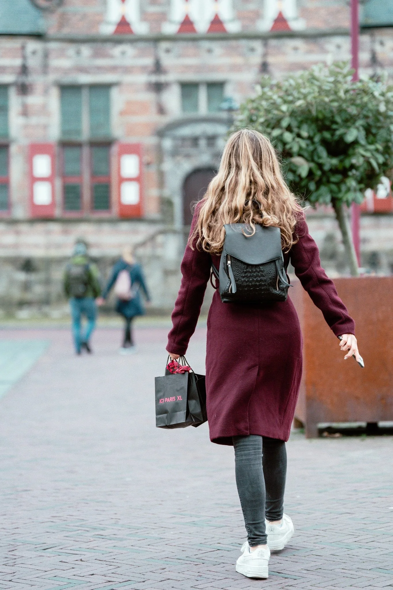 Vrouw met lang haar in een vin rouge jas, wandelen op straat met een boodschappentas en rugzak, achtergrond van een oude gebouw en een boom.