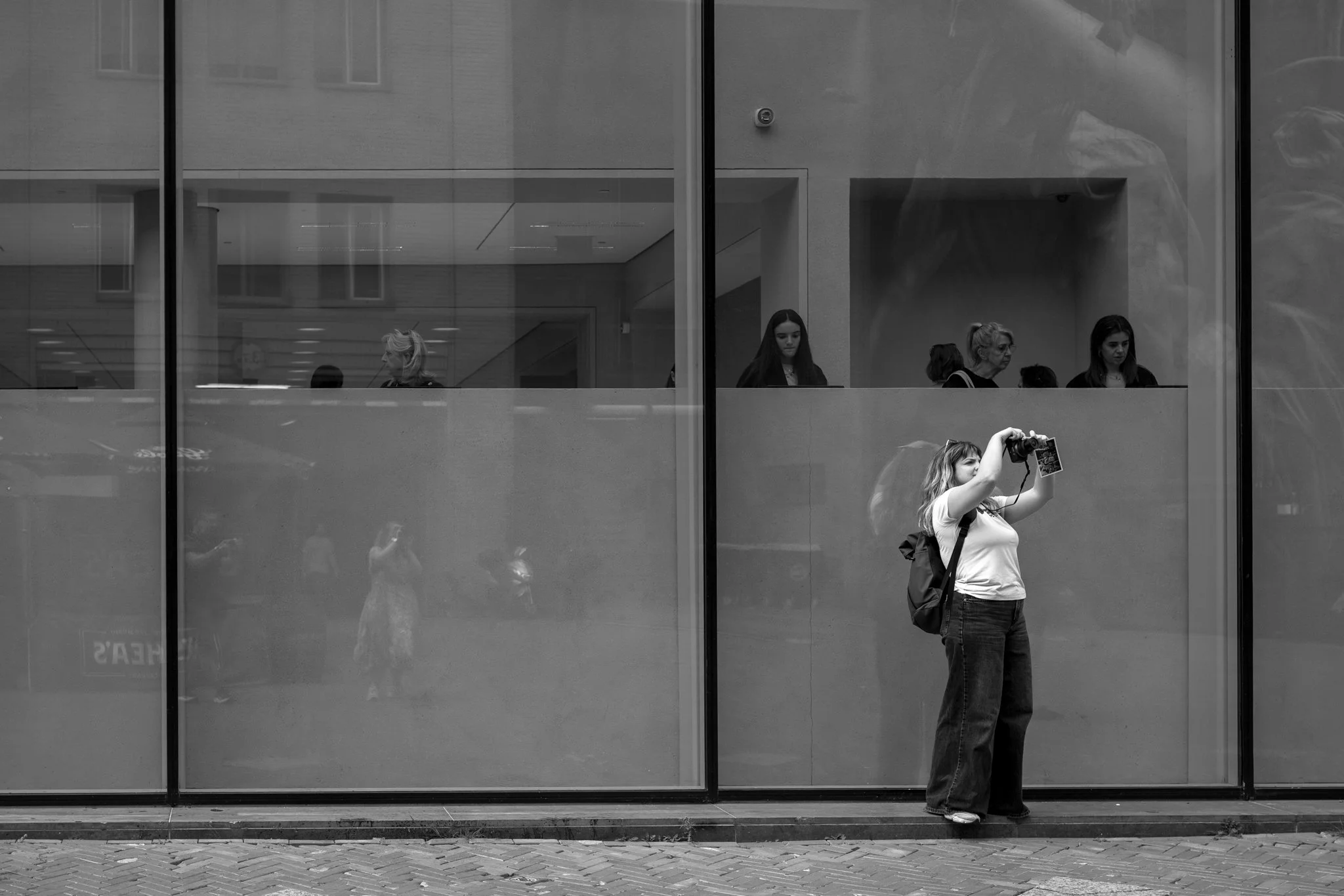 Een vrouw met een rugzak die een camera vasthoudt en aan het fotograferen is voor een groot raam, met mensen binnen die door het raam zichtbaar zijn.