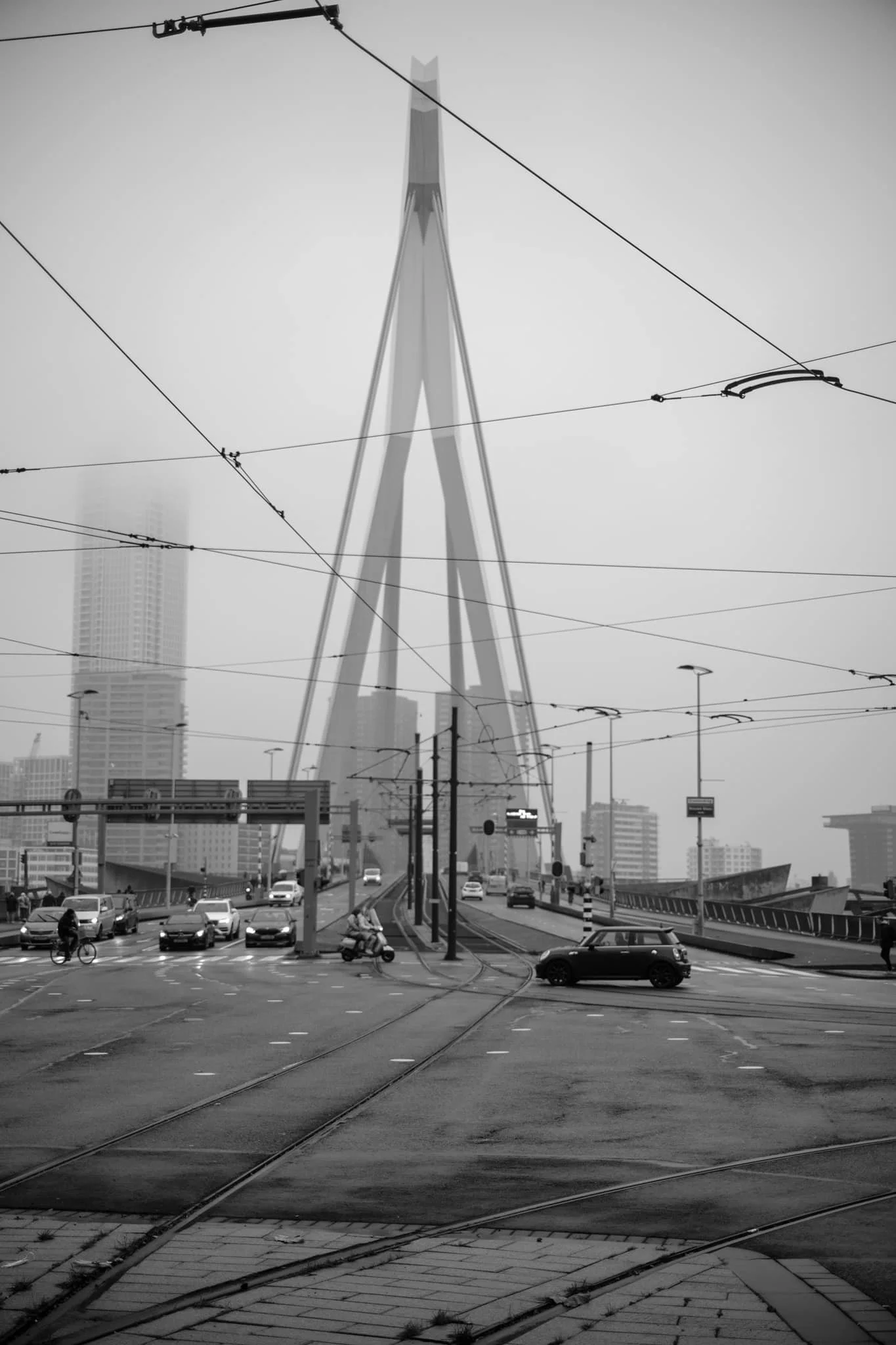 Een zwart-wit foto van een stadsgezicht met een grote futuristische toren op de achtergrond, een weg met tramlijnen, voertuigen, fietsen en mensen.