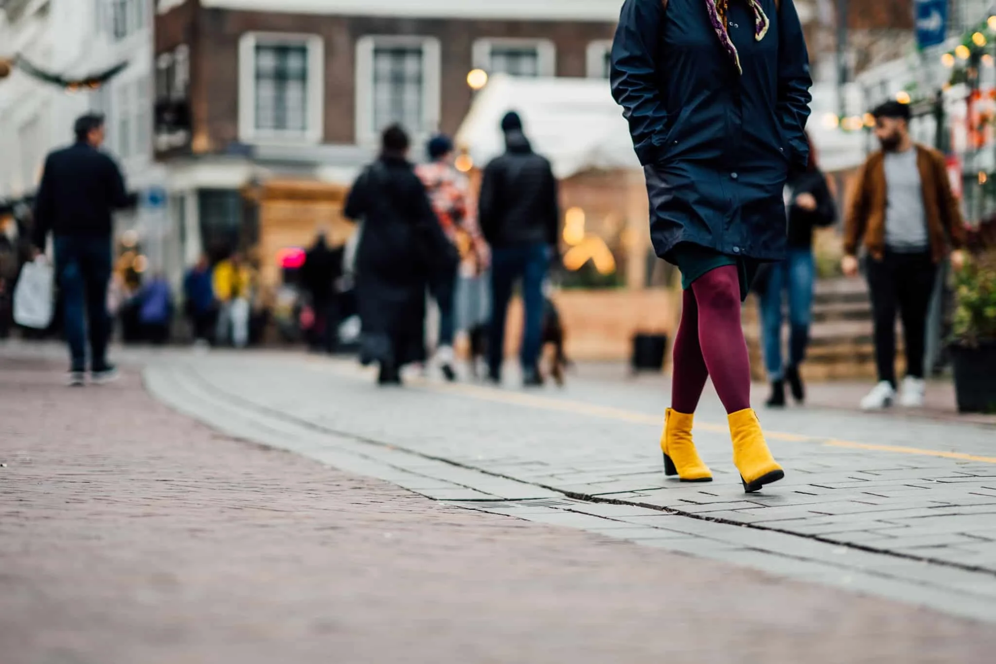 Close-up van een persoon die op hoge gele schoenen en die burgundy legging lopen, terwijl andere mensen op de achtergrond op straat lopen in een stad, wat lijkt op een reünie of markt.