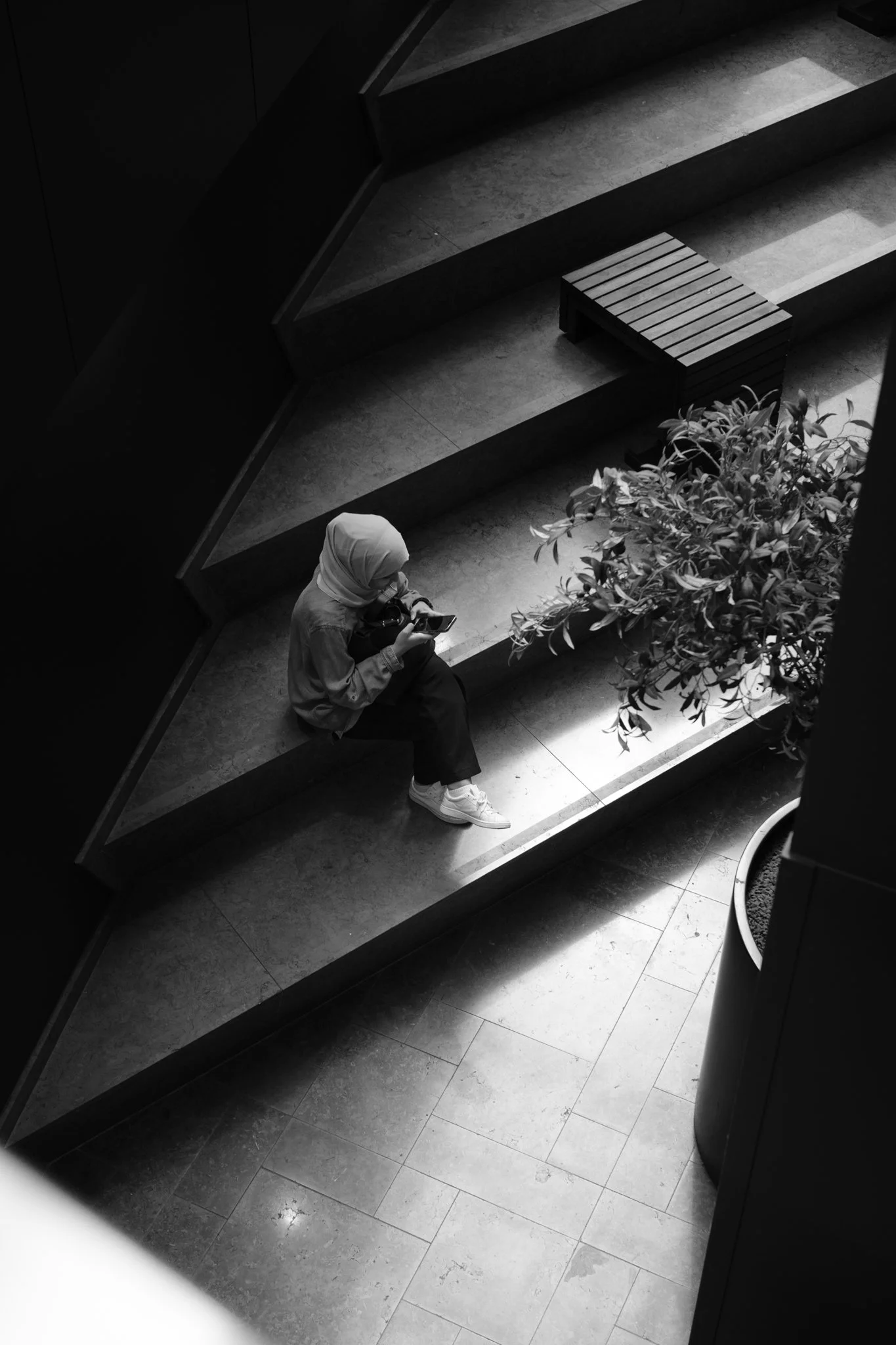 Een vrouw met een hoofddoek zit op een betonnen bankje, verdiept in haar telefoon, naast een grote plant, in een modern gebouw.