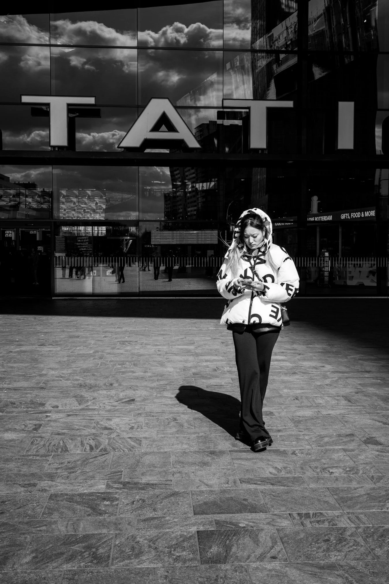 Een vrouw die buiten een modern glazen gebouw staat, gekleed in een pomponjack en zwarte broek, terwijl ze op haar telefoon kijkt.