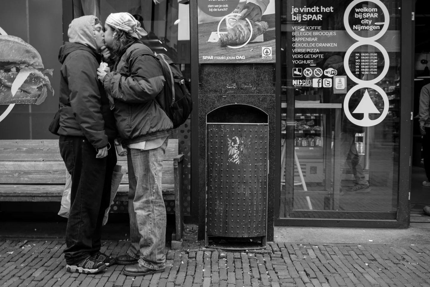 Twee mensen kussen elkaar voor een supermarkt in Nijmegen. Ze dragen jassen en hoeden, en staan op een bakstenen stoep.