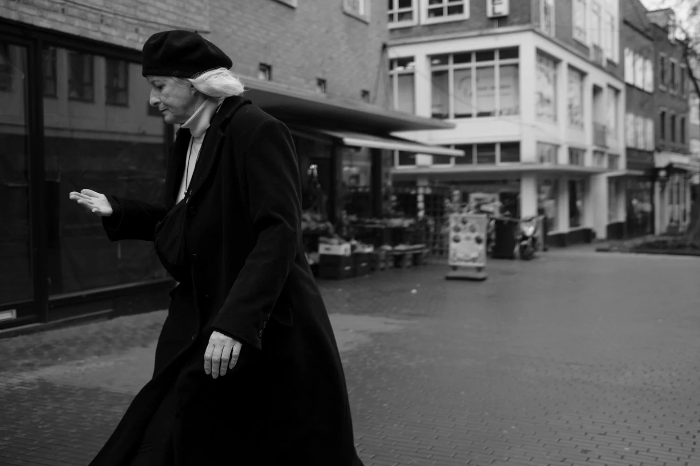 Een oudere vrouw loopt een straat op, gekleed in een lange jas en een hoofddoek, in een stedelijke omgeving met moderne gebouwen.