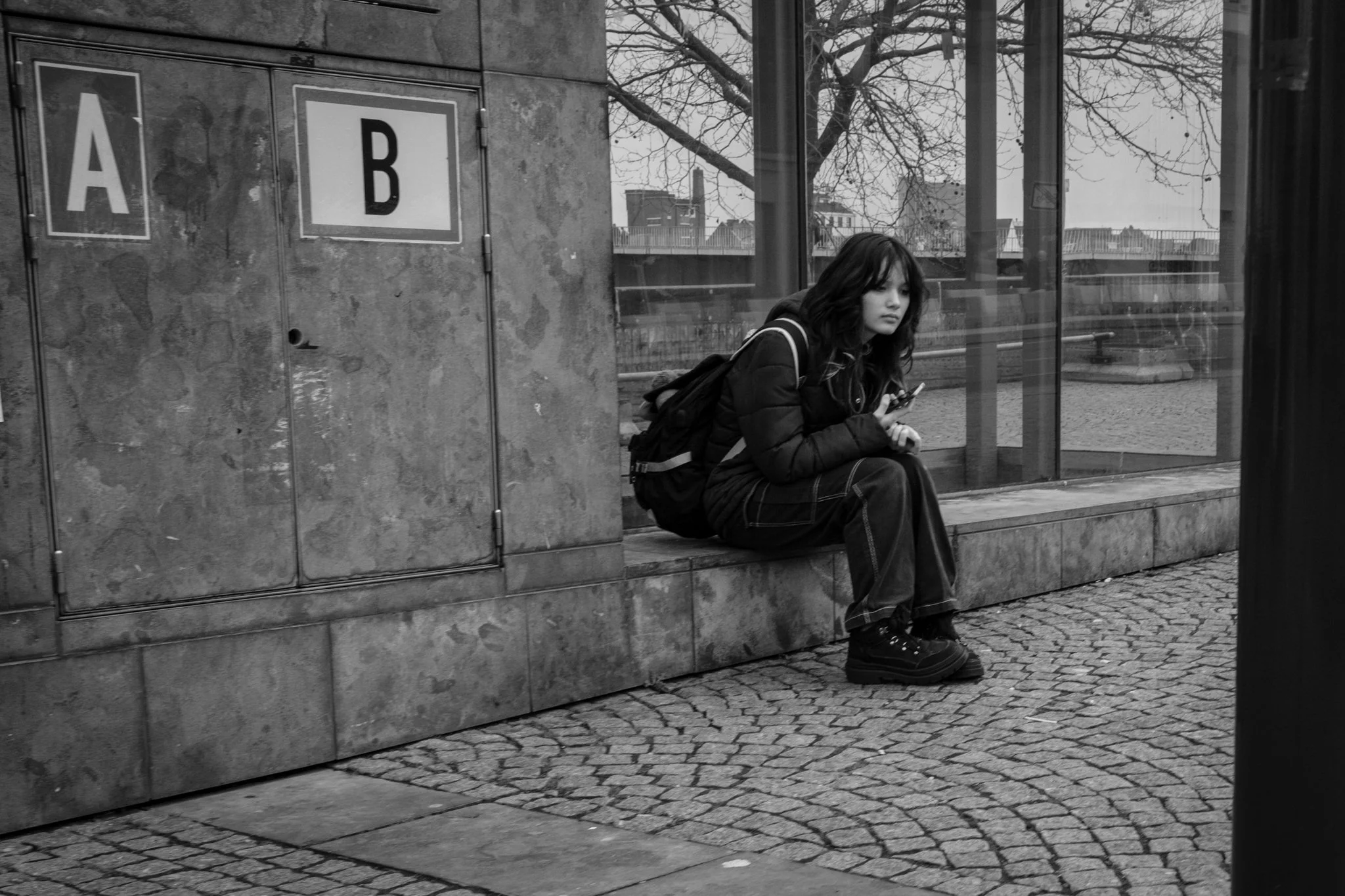 Een jonge vrouw met een rugzak zit op een bankje, ze kijkt naar haar telefoon en is gekleed in een donkere jas en schoenen, achter haar grote ramen zijn bladeren van een boom zichtbaar.