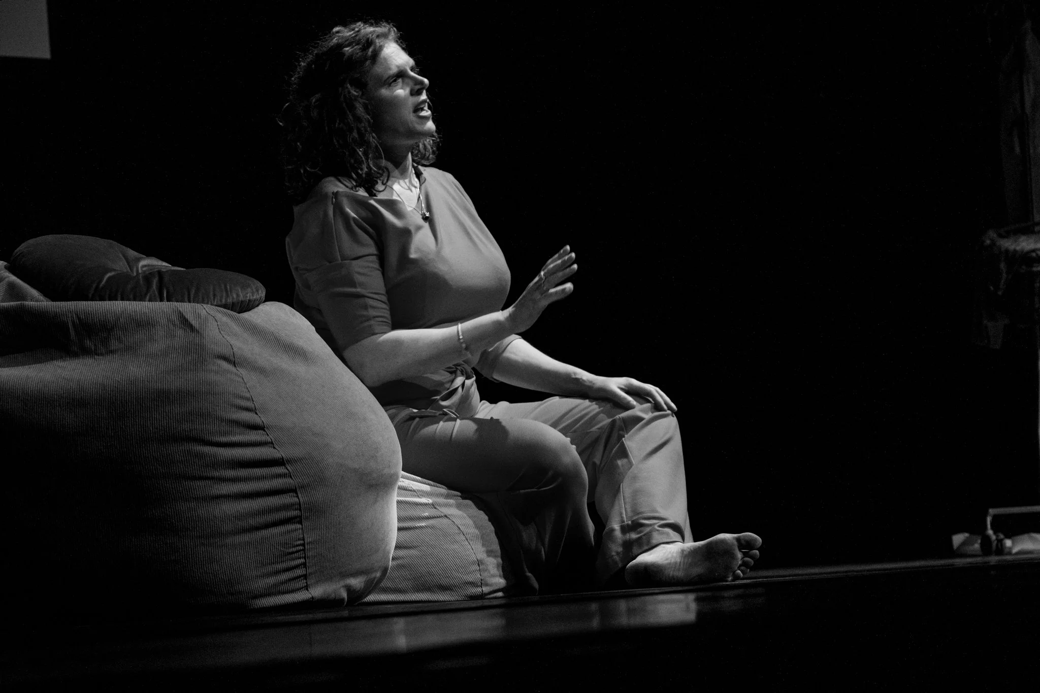 Een vrouw zit in het theater op het podium op een zitkussen, haar hand omhoog en druk vertellend.