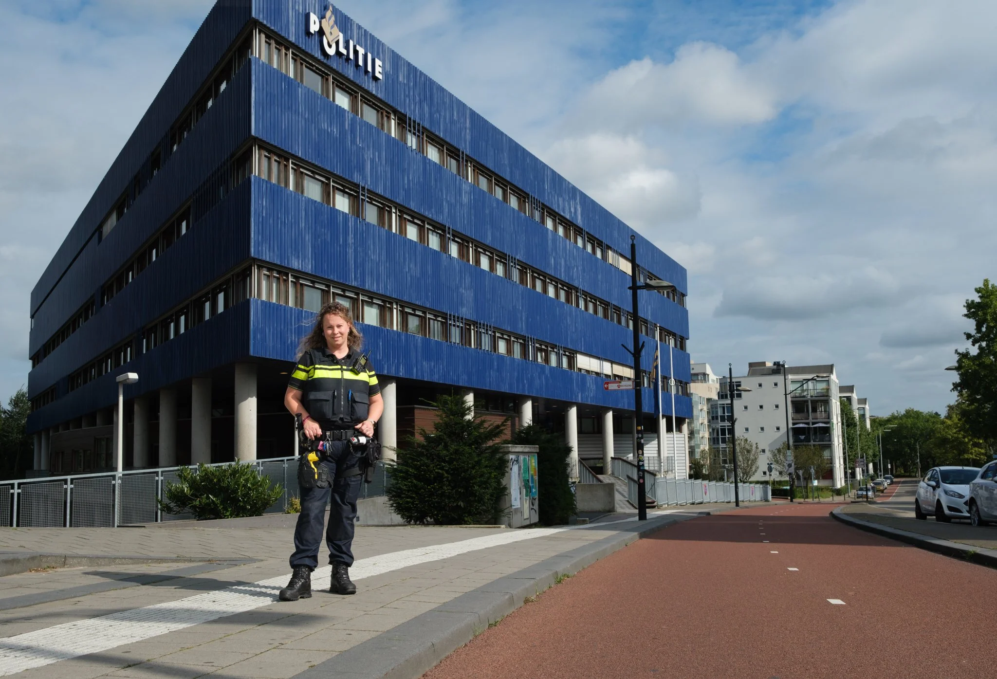 Een politieagent staat op een zebrapad voor een modern blauw kantoorgebouw in een stad, met auto's op de weg en een bewolkte hemel.