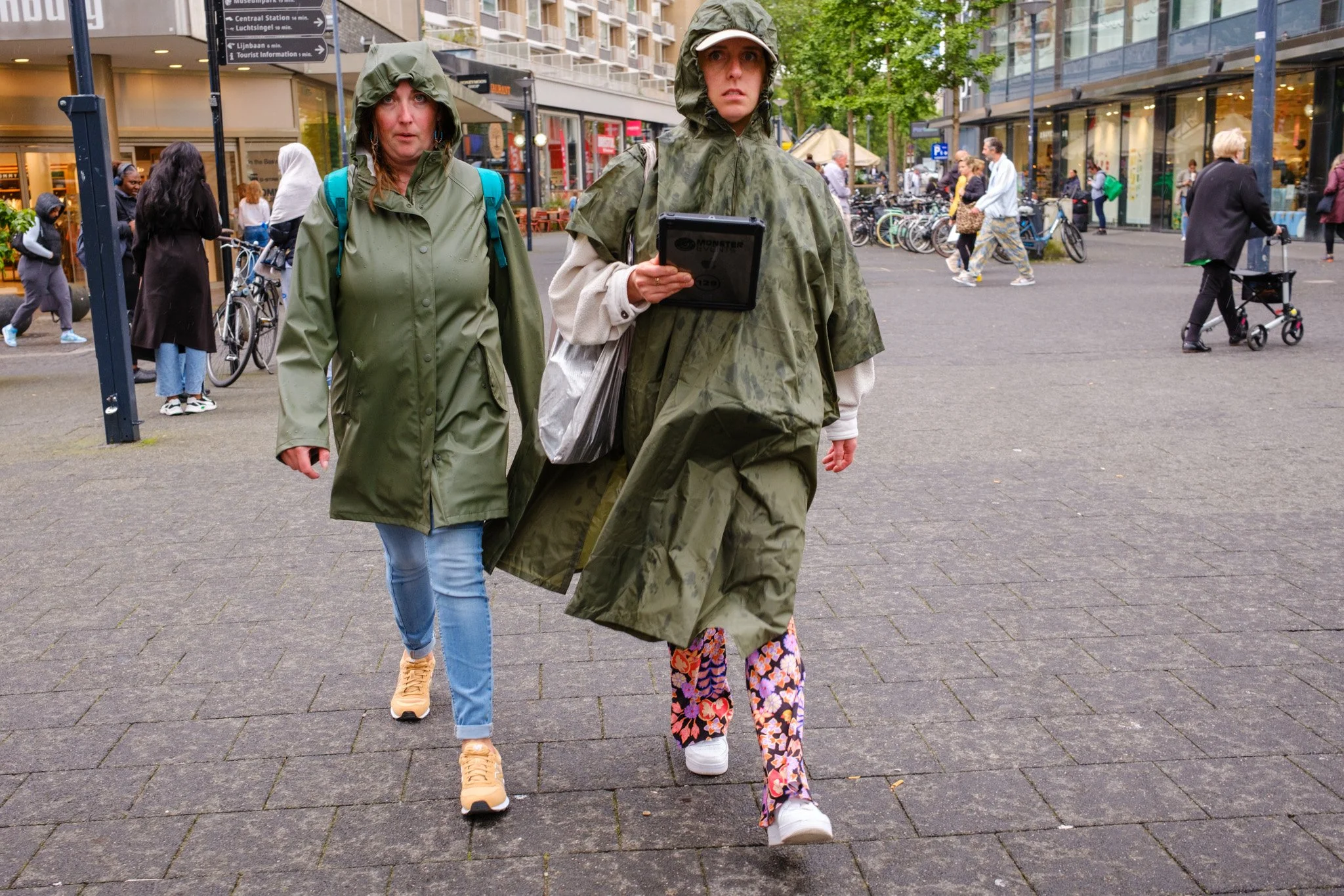 Twee mensen in regenjassen lopen op een stadsplein, omringd door andere voetgangers, fietsen en winkels.