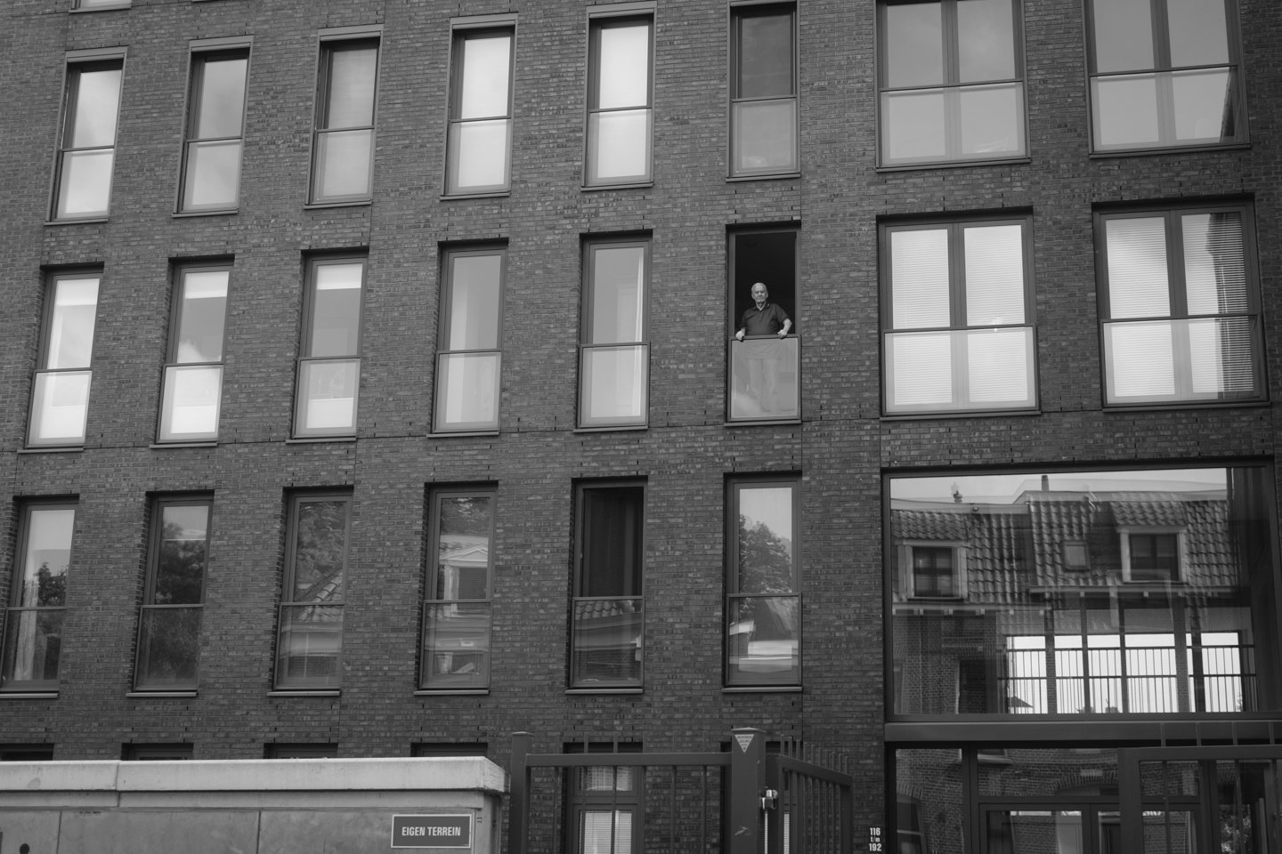 Een man staat op een balkon van een modern bakstenen appartementengebouw met grote ramen.