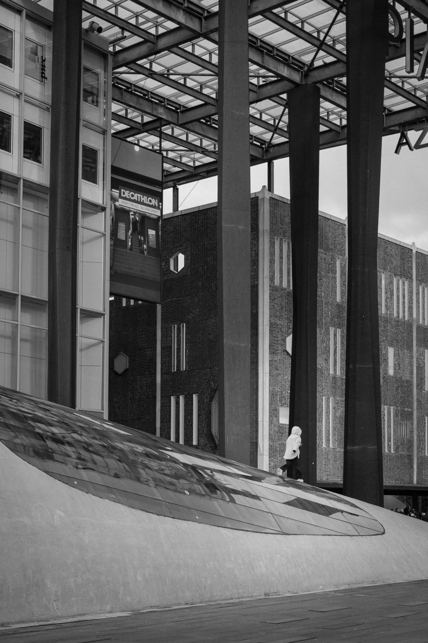 Een zwart-witfoto van een stedelijk gebied met moderne architectuur, glazen gevels, grote stevige metalen pilaren, en een persoon in een witte jas die schaatst of glijdt over een lage, gladde structuur. Op een digitaal scherm is een advertentie voor 