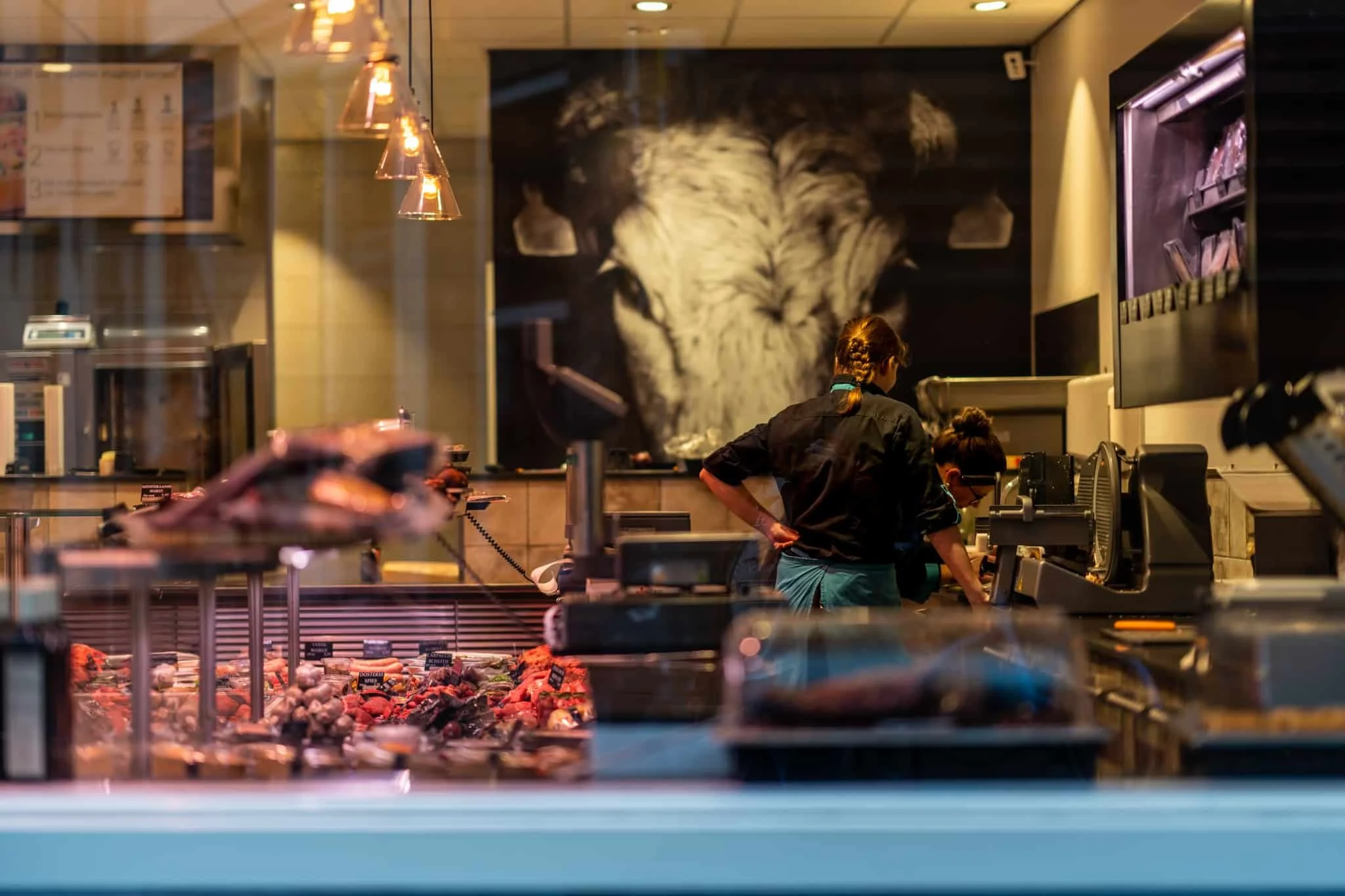 Twee vrouwen werken in een vleeswinkel met een uitstraling van een moderne, sfeervolle omgeving. In de voorgrond liggen verschillende stukken vlees uitgestald, terwijl de vrouwen achter de toonbank bezig zijn.