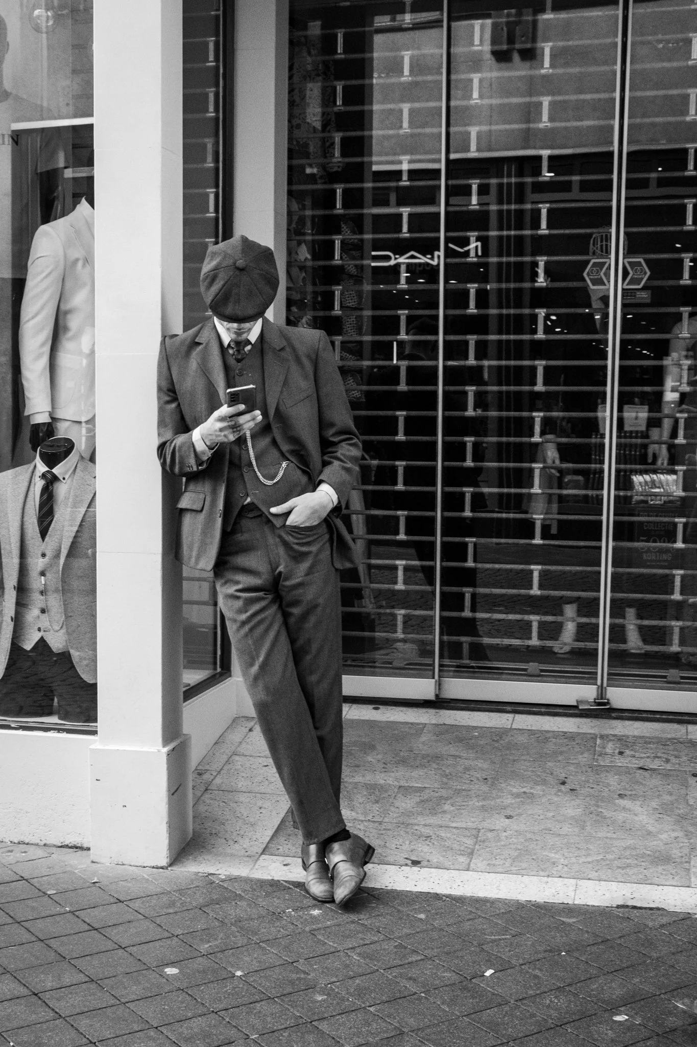 Zwarte en witte foto van een man in een zwart pak en hoed die op een straat zit en op zijn telefoon kijkt voor een kledingwinkel.