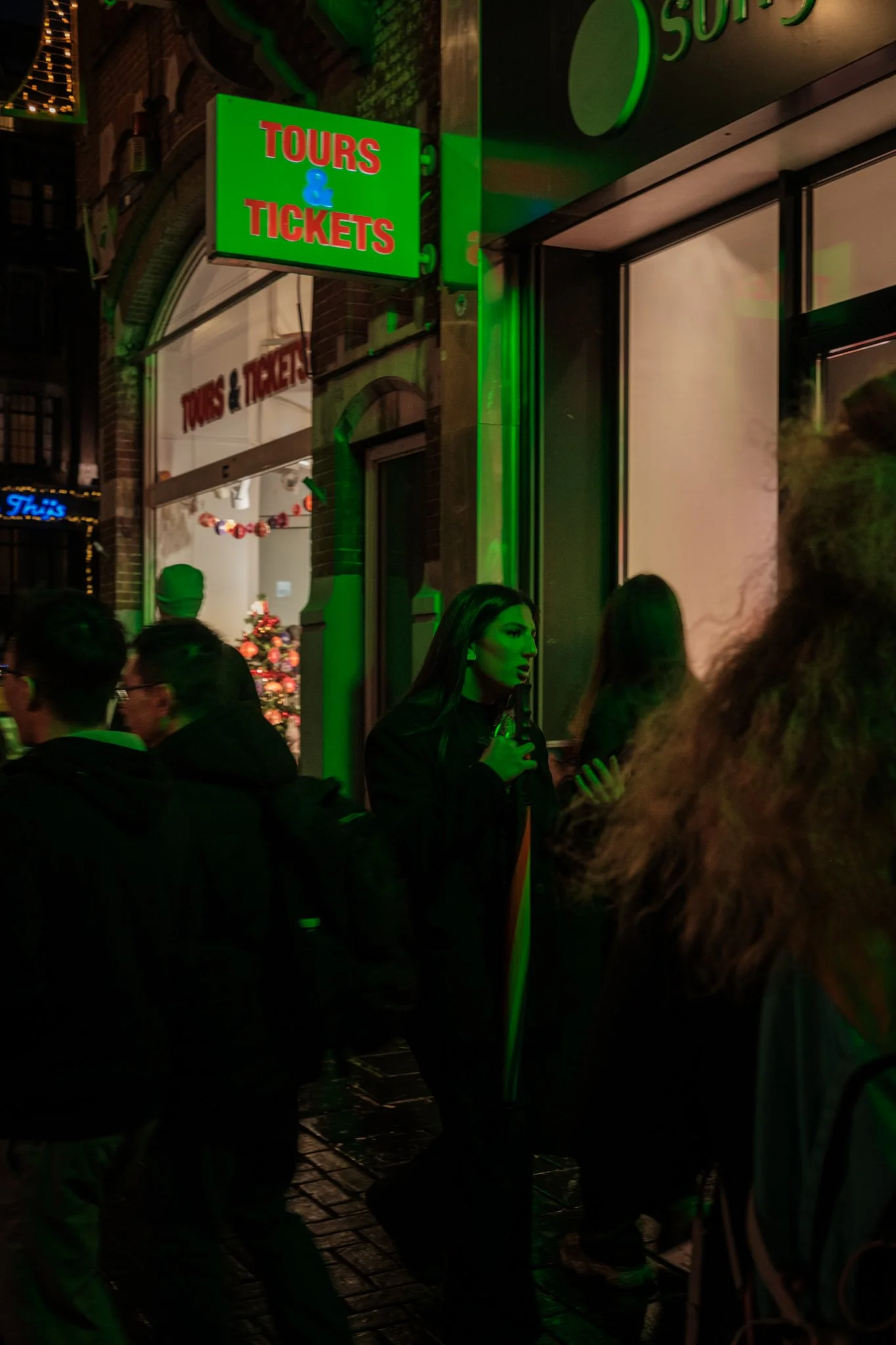 Groene neonbord met de tekst 'TOURS & TICKETS' buiten een gebouw tijdens de avond. Mensen wachten in de rij voor een gokhal met een kerstboom zichtbaar door de deur.