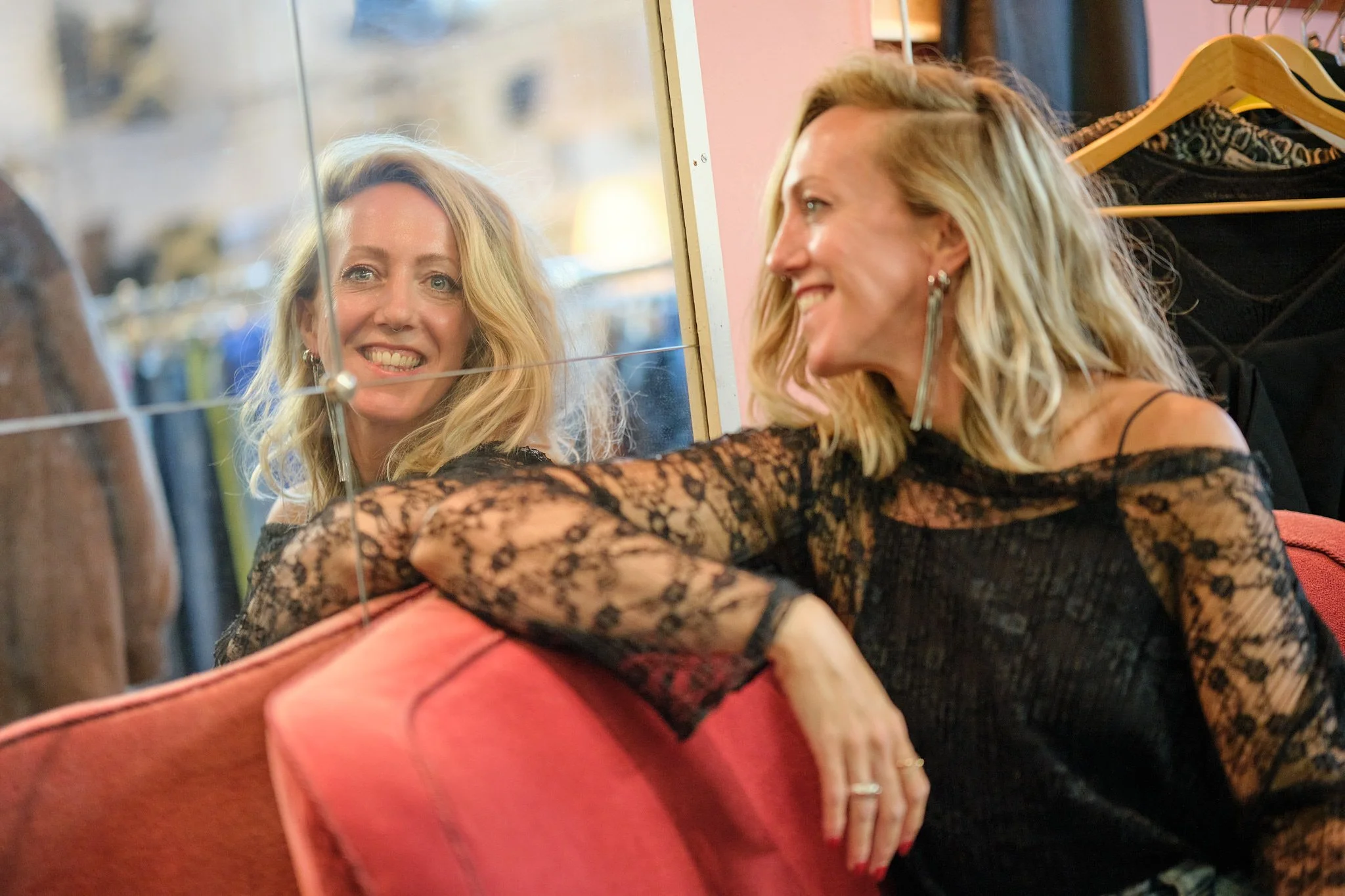 Een vrouw met blond haar, gekleed in een zwart kanten shirt, kijkt in een spiegel en glimlacht.