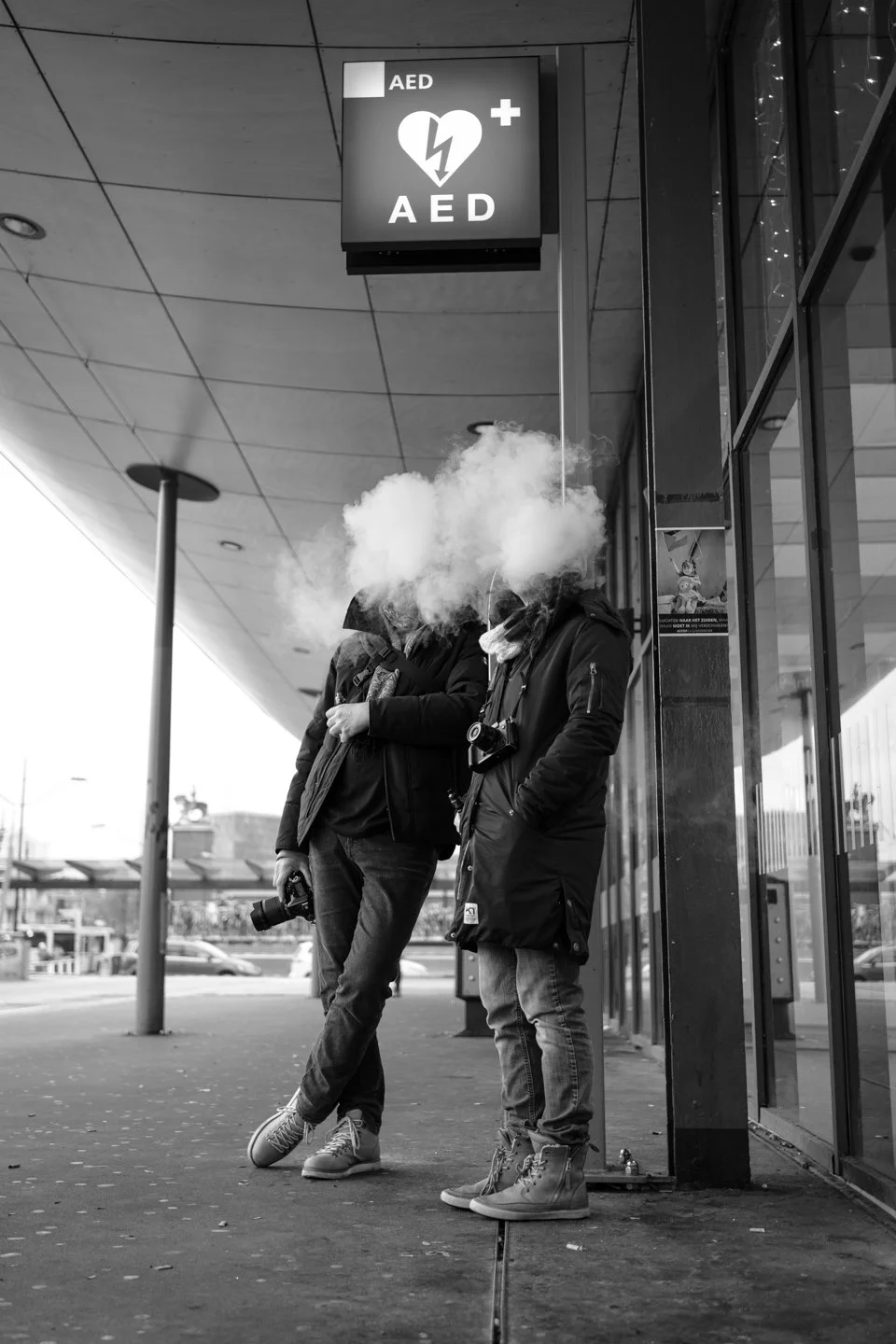 Twee mensen staan buiten bij een gebouw, beide met hun gezichten verstopt achter een wolk van rook of damp. Een persoon houdt een camera, de ander draagt een tas en een zaklamp. Boven hen hangt een rood bord met witte tekst 'AED' en een hartslaglijn.