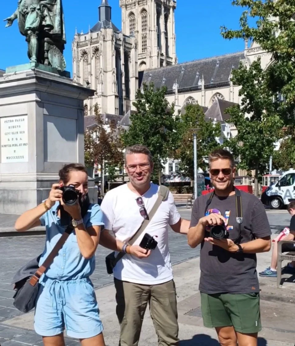 Drie mensen met camera's staan voor de kathedraal van Antwerpen. De torens en het glas-in-loodraam van de kathedraal zijn zichtbaar op de achtergrond.