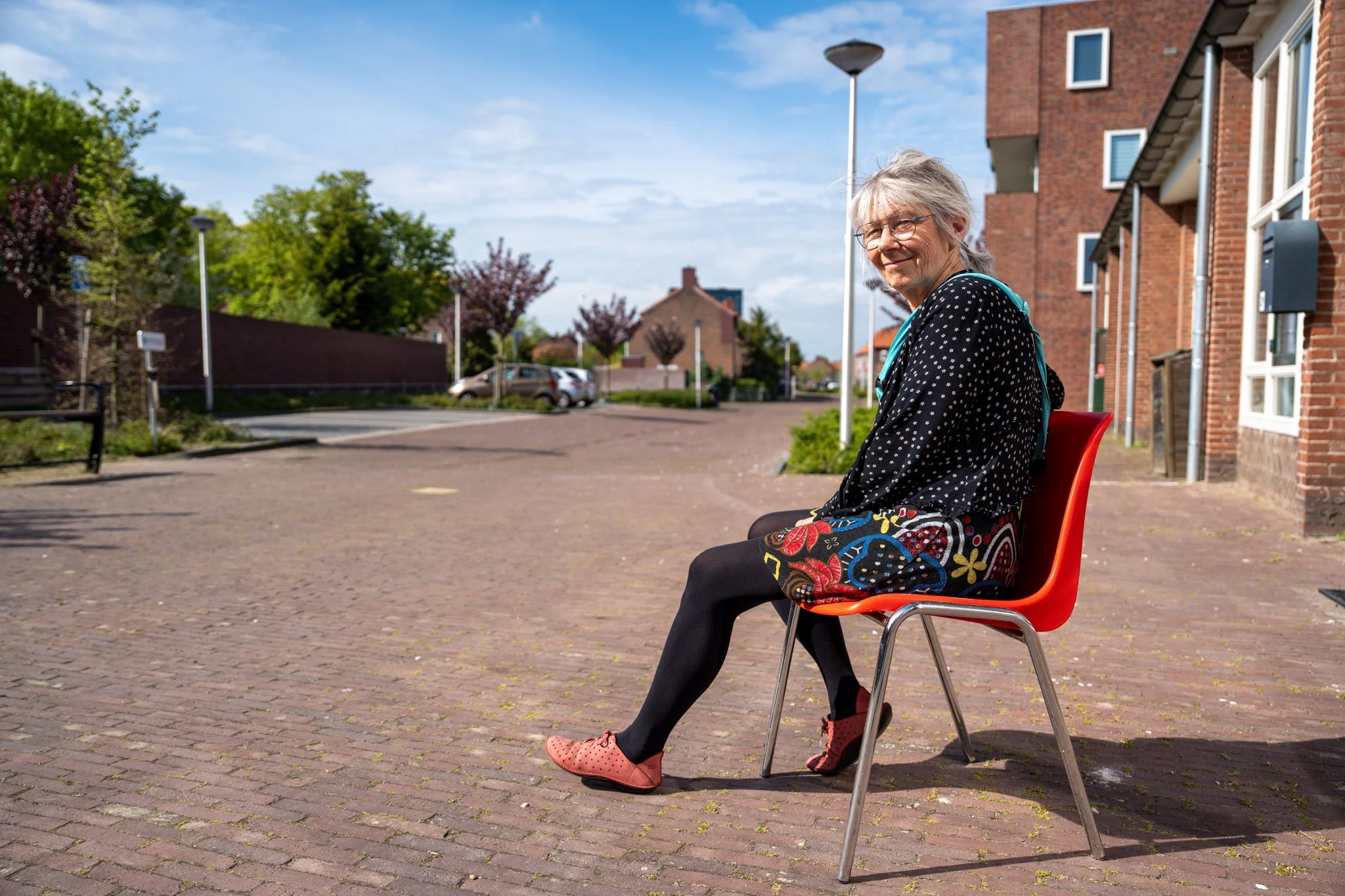 Een vrouw zit op een rode plastic stoel op een straat met gebakken klinkers. Ze draagt een zwarte jurk met kleurrijke patronen, een zwarte jas met witte stippen, zwarte panty en zolen in roze. Ze heeft grijs haar en draagt een bril. Op de achtergrond