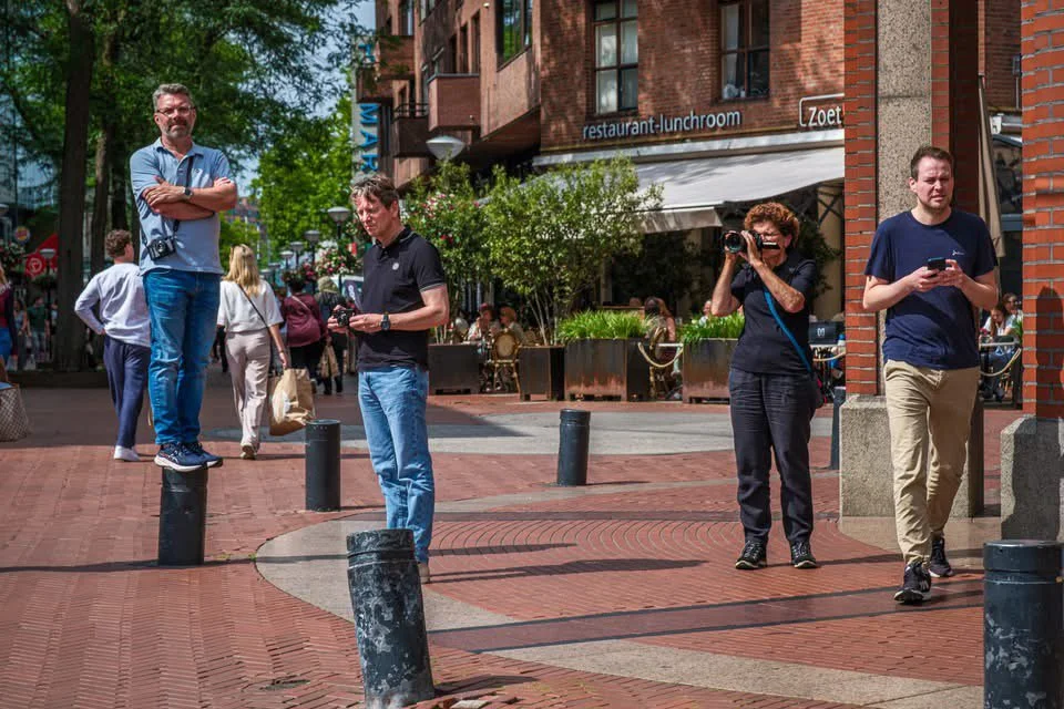 Vier mensen op een stadsplein, een man staat op een paal, een vrouw met een camera, twee mannen die op hun telefoons kijken, in de achtergrond mensen, bomen, en een restaurant.
