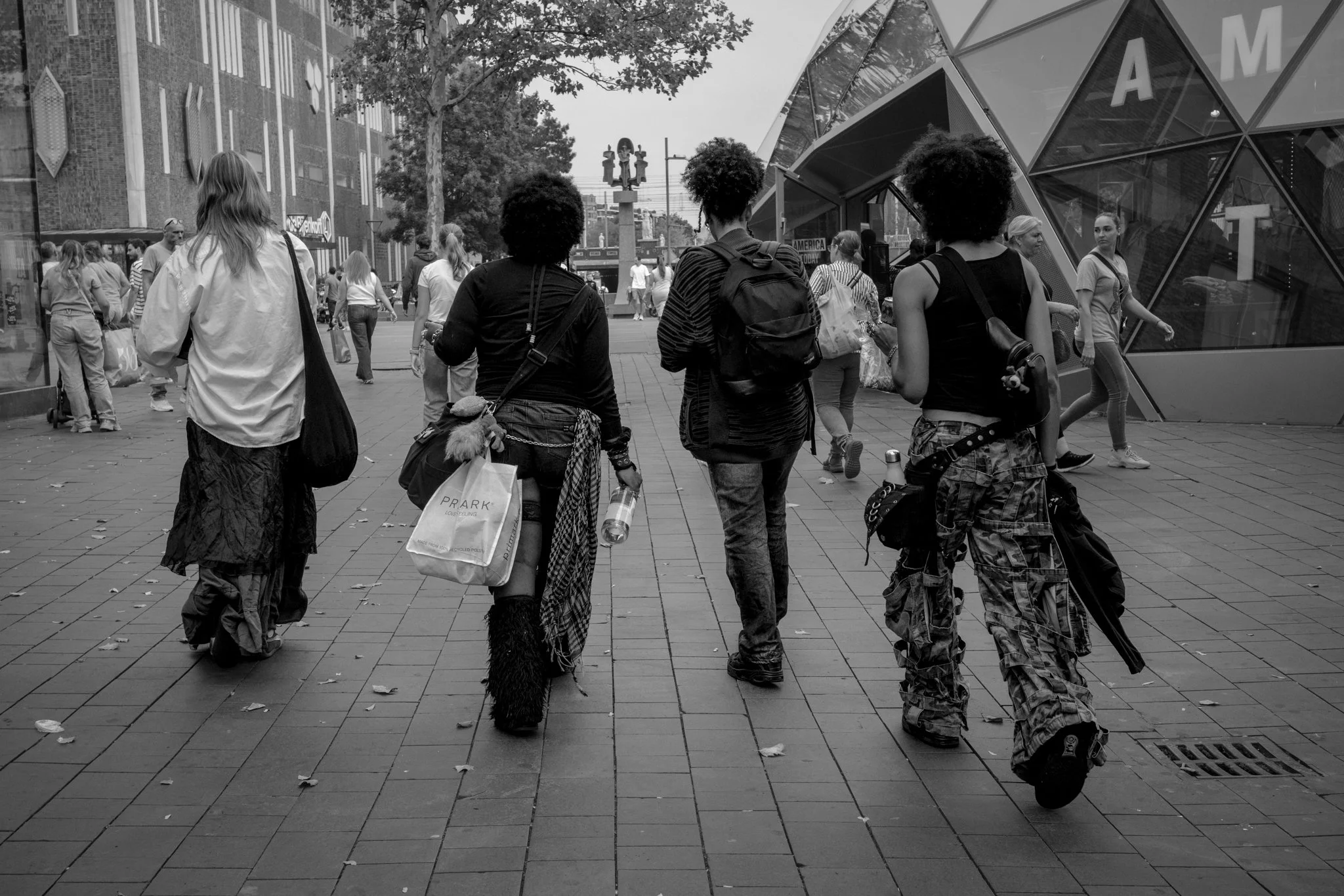Vijf straatverkoopsters lopen door een stad, met verschillende shoppingtassen en spullen. Andere voetgangers in de achtergrond, moderne gebouwen en bomen.