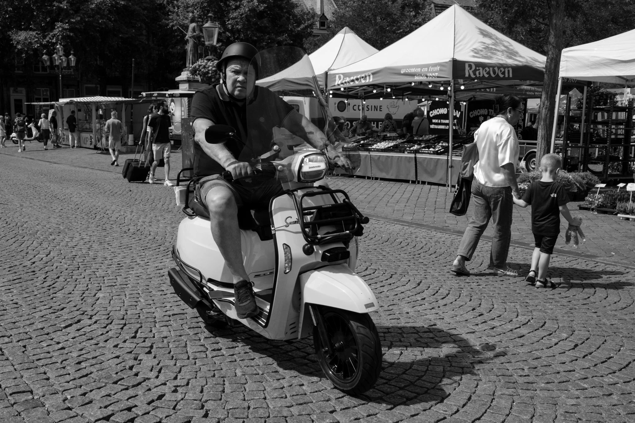 Man rijdt op een elektrische scooter op een marktplein met kraampjes en mensen die winkelen en wandelen.