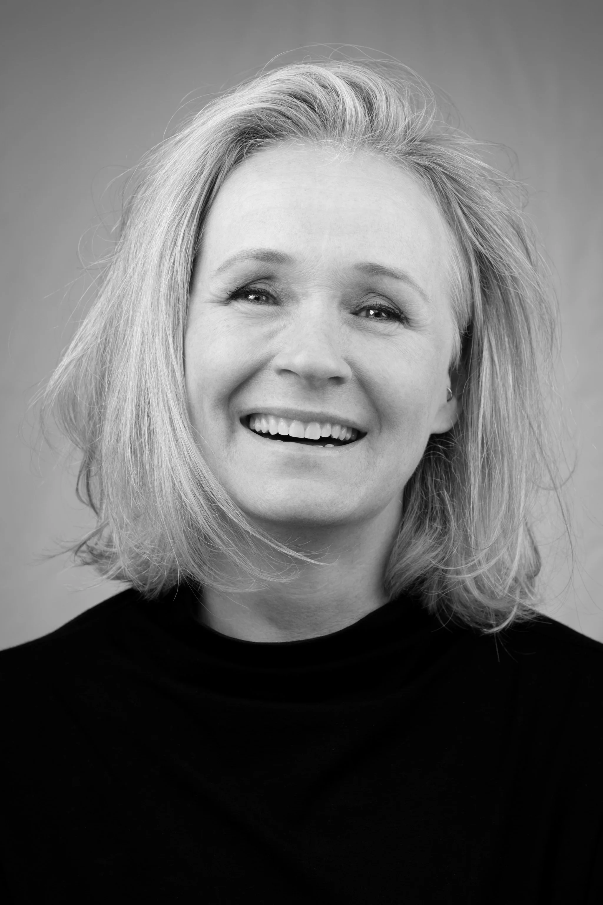 Zwart-wit portret van een glimlachende vrouw met schouderlange, golvende haar, gekleed in een zwarte top.