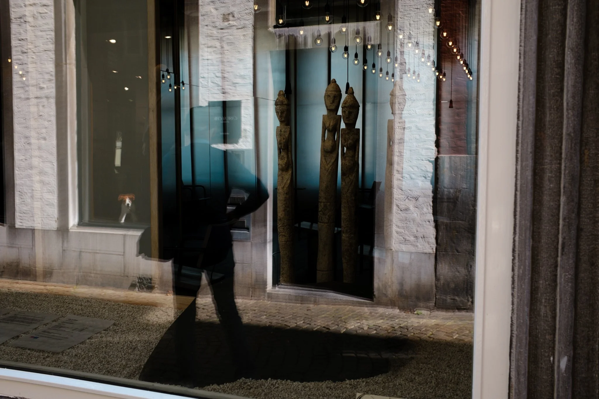 Reflectie van een persoon en een hond in een raam, met treden en beeldjes van mensen in de etalage.