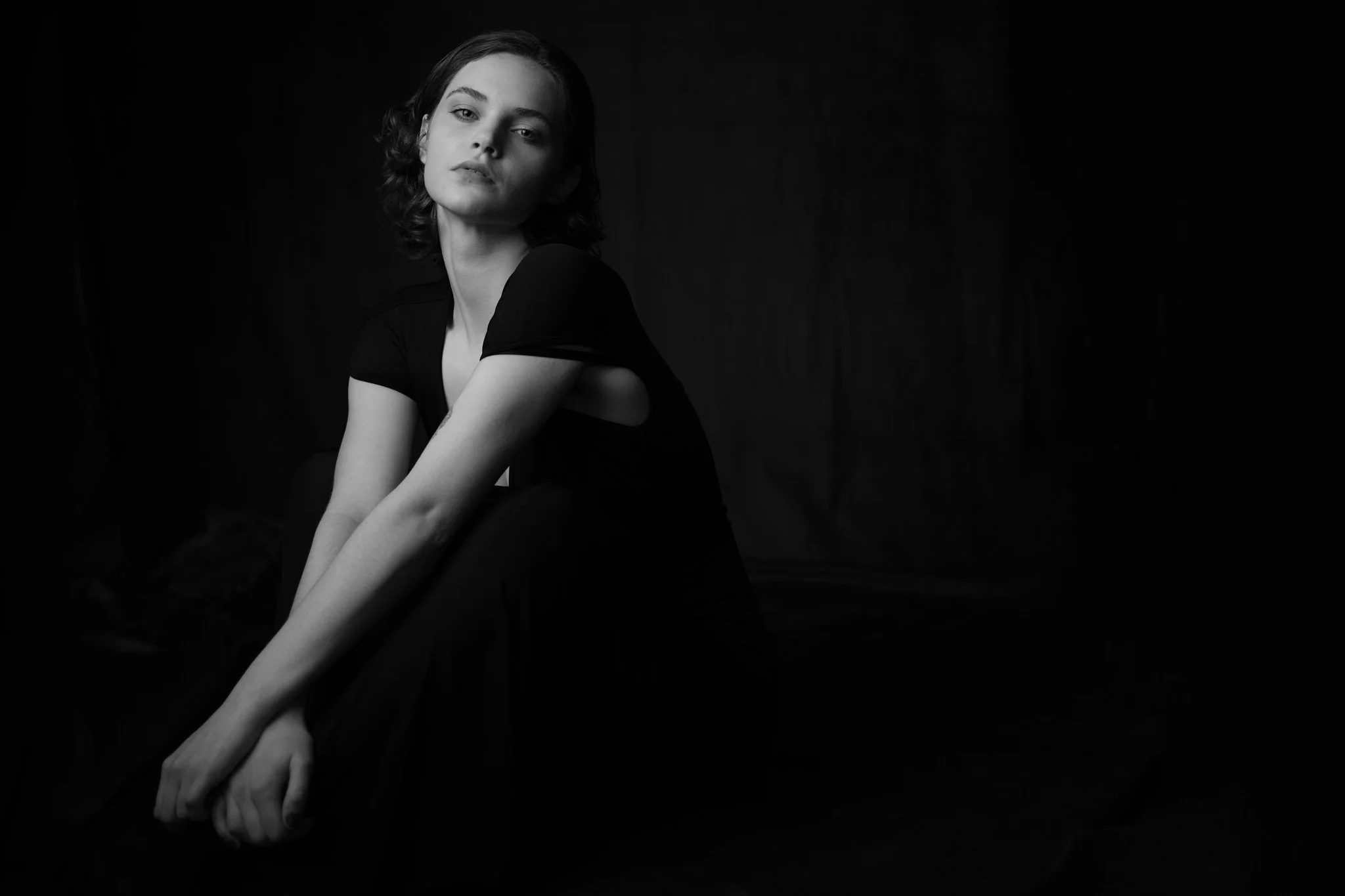 Zwarte en witte foto van een jonge vrouw met kort, golvend haar, zittend met haar benen ingekruist, kijkend naar de camera, met een donkere achtergrond.