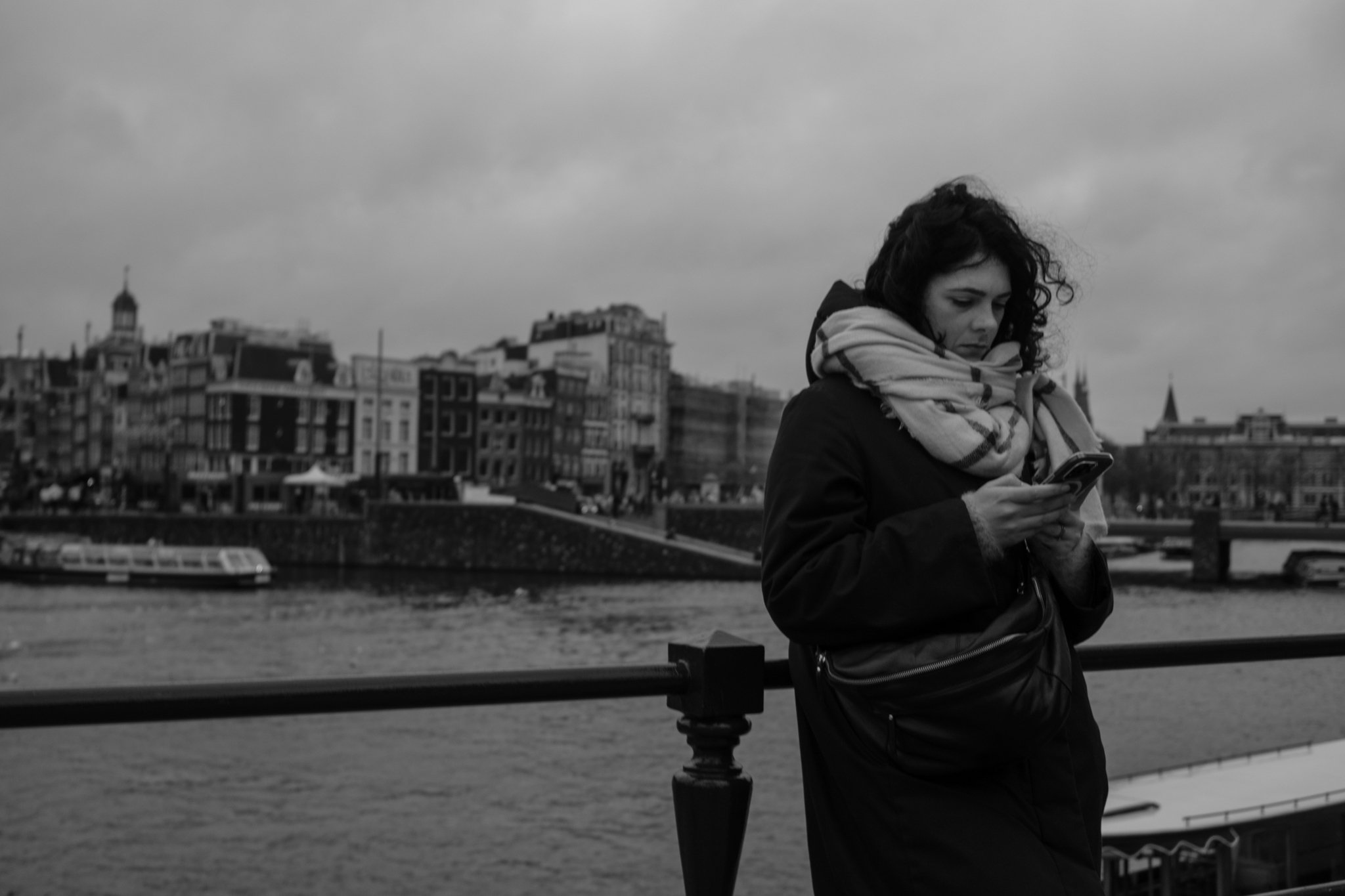 Een vrouw met een grote scarf kijkt op haar smartphone tegen een achtergrond van een rivier en gebouwen in een stad, mogelijk Parijs, op een bewolkte dag.