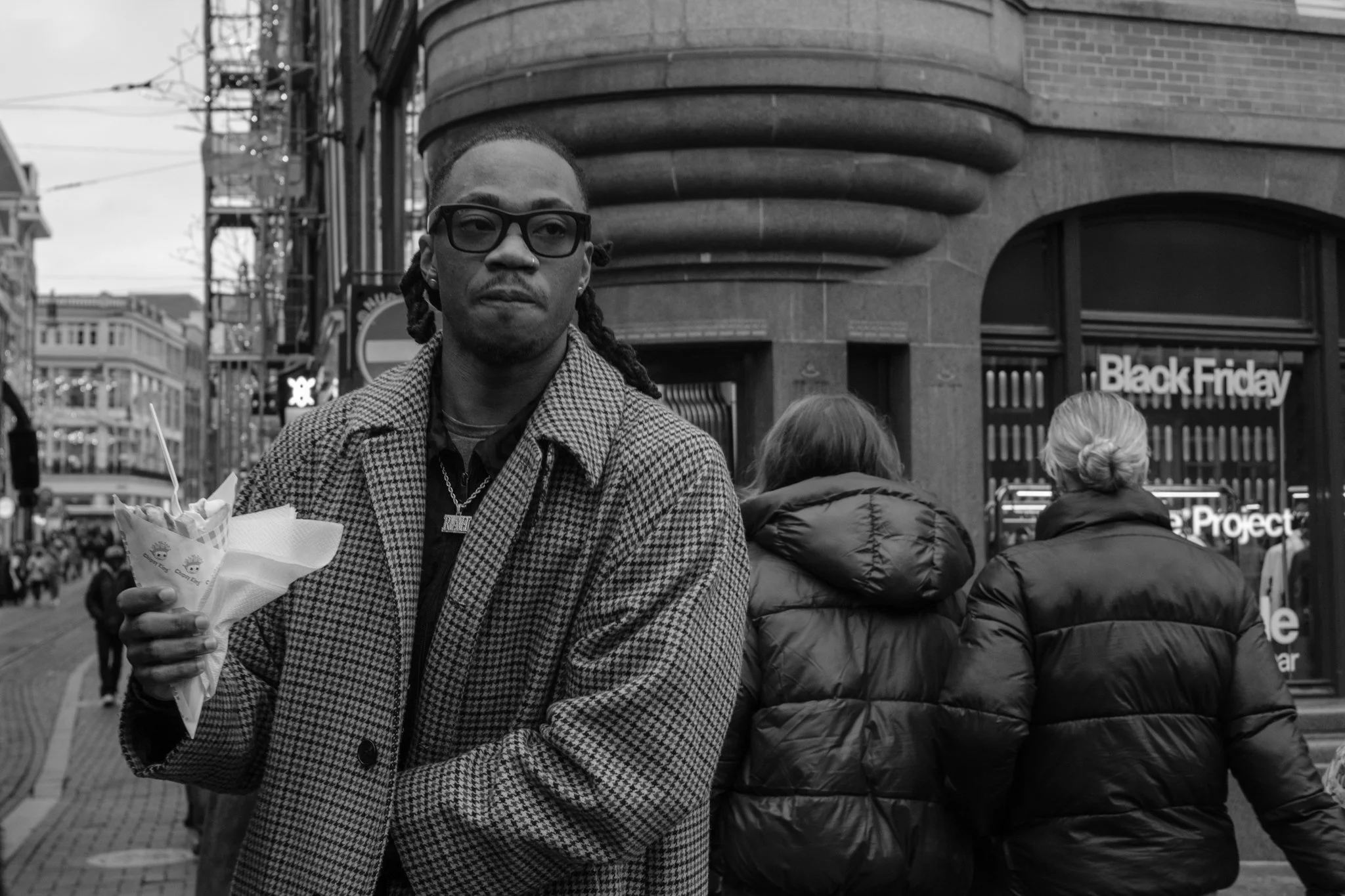 Een man met lang haar en een spijkerjack houdt een ijsje in zijn hand, staande op een straat in een stadscentrum. Op de achtergrond zijn drie mensen met winterjassen te zien en een winkel met een bord dat 'Black Friday' zegt.