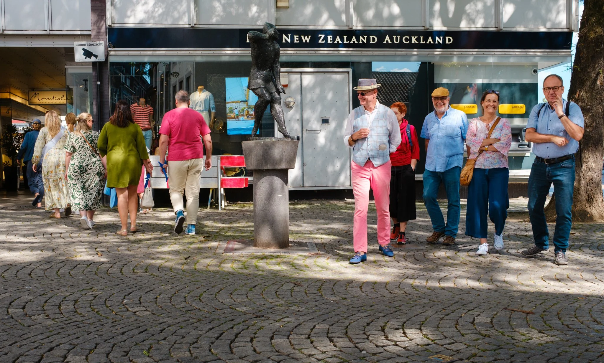 Mensen wandelen op een plein met kasseien, een standbeeld en winkels op de achtergrond.