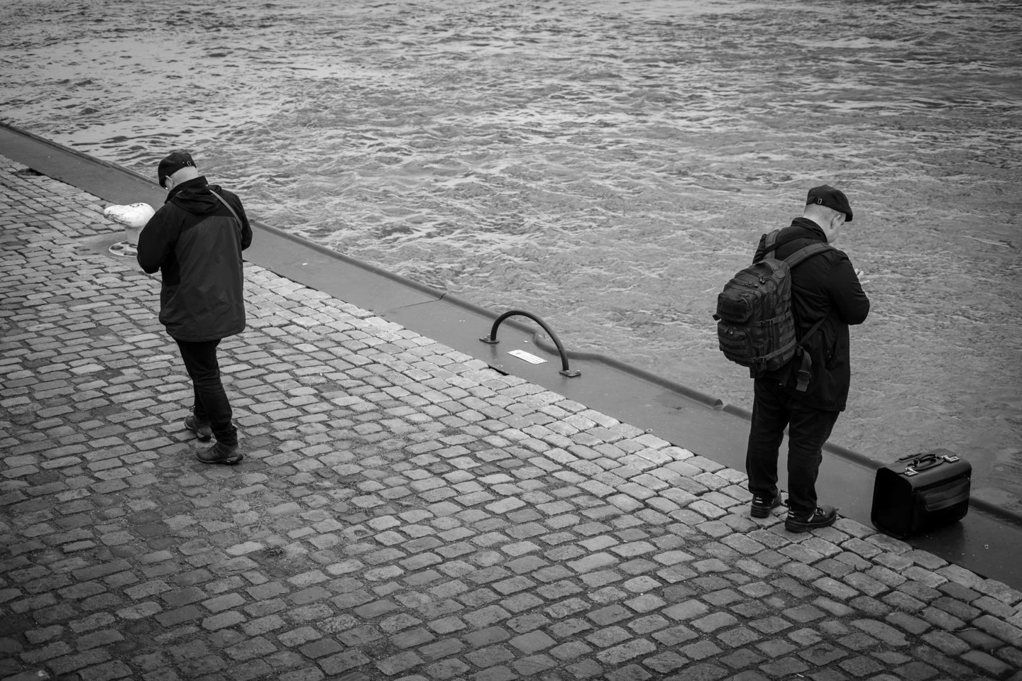 Twee mannen staan aan de waterkant, één met een grote rugzak en de ander met een kleine koffer en een tas, terwijl ze naar het water kijken op een kade met stenen geplaveid.