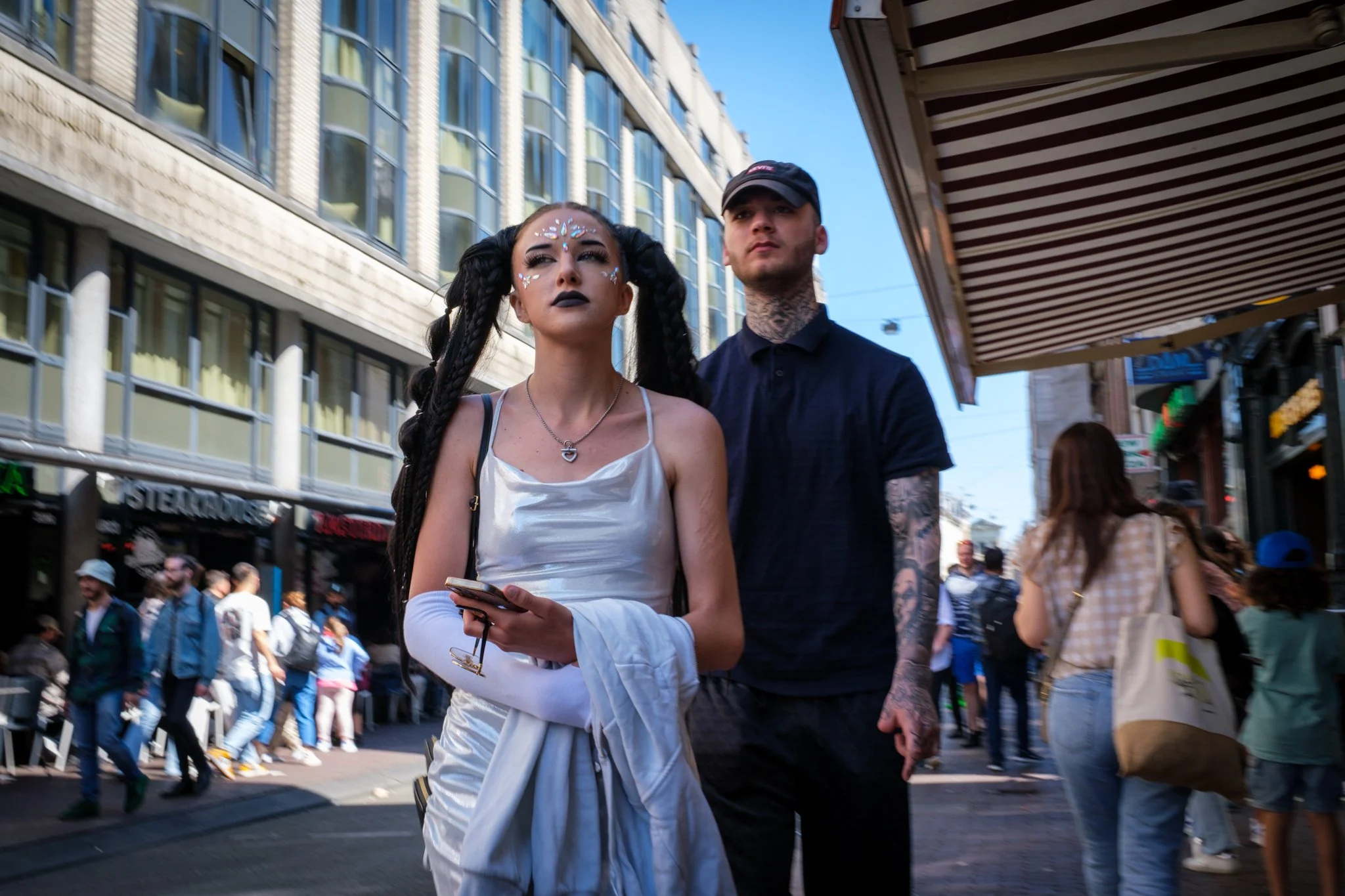 Een jonge vrouw met opvallende make-up en vlechtkapsels staat op een drukke stadss straat, naast een man met tattoos, beide kijken vooruit.