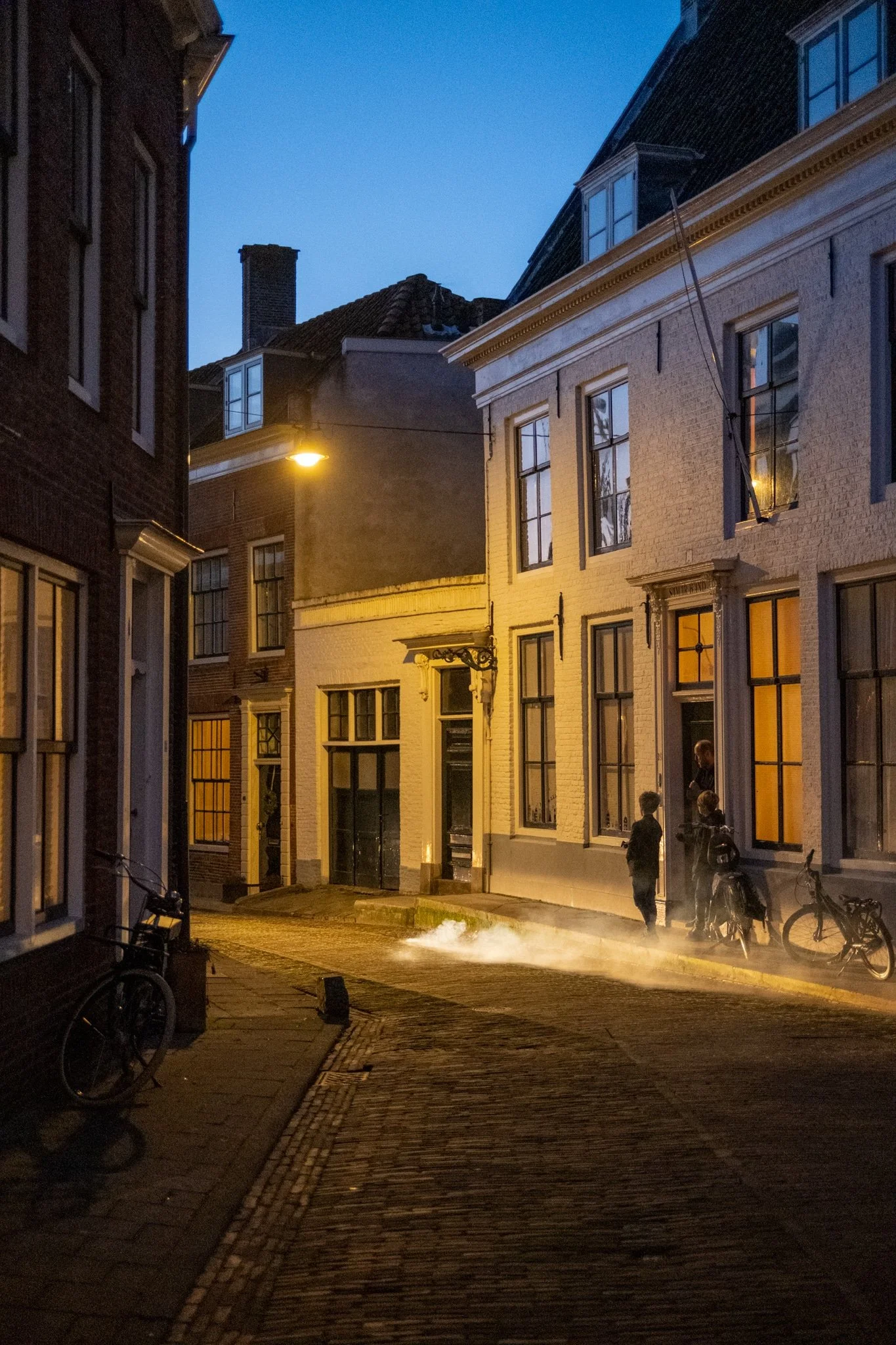 Een straat in de avond met ouderwetse gebouwen, fietsen op de stoep, een straatlantaarn en mensen die praten voor een wit pand. Er is wat stoom of rook op de straat.