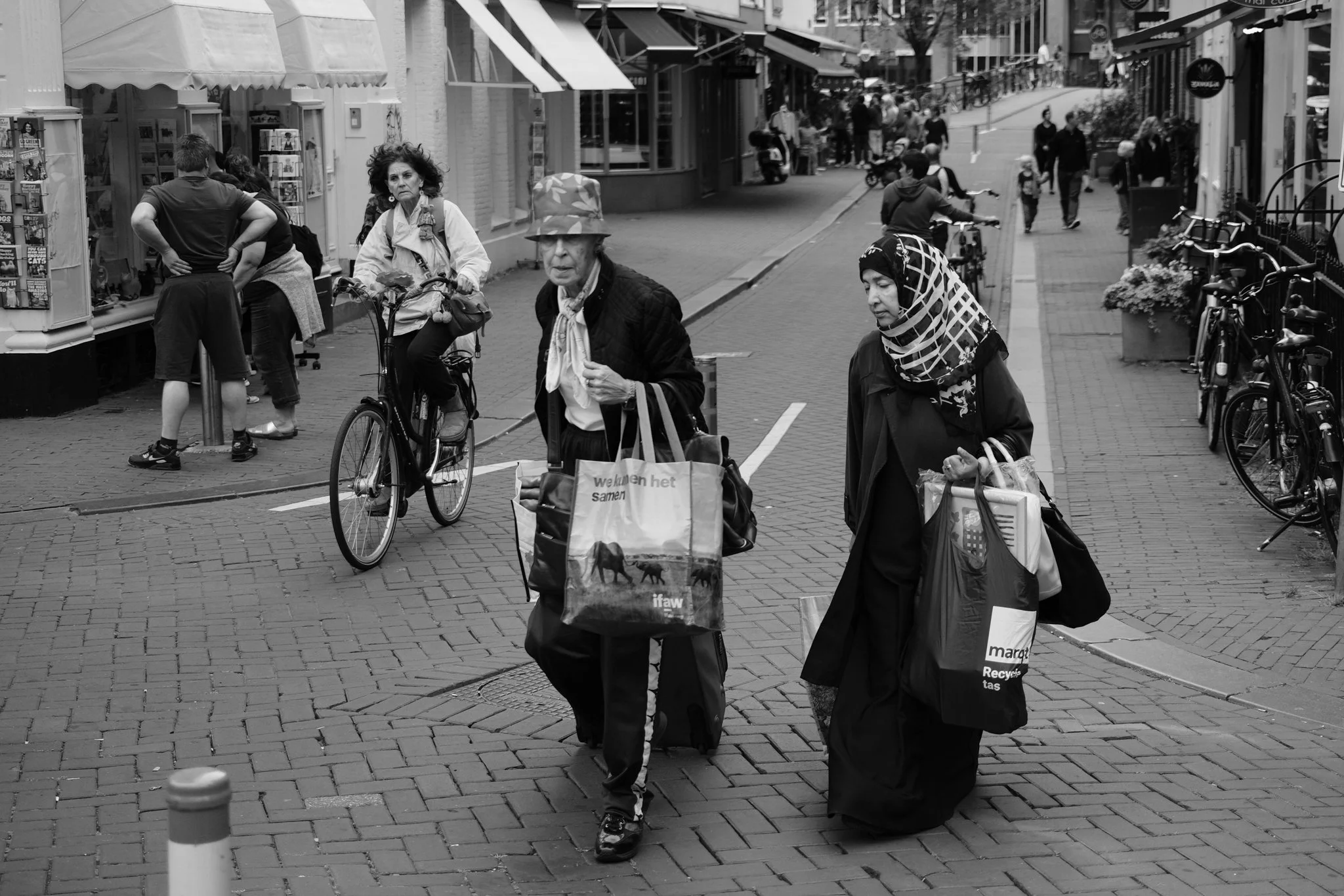 Twee vrouwen lopen op een straat met winkelgebieden, een vrouw op hakken met een grote tas en een vrouw met een hoofddoek met meerdere tassen, terwijl mensen op de achtergrond fietsen en winkelen.