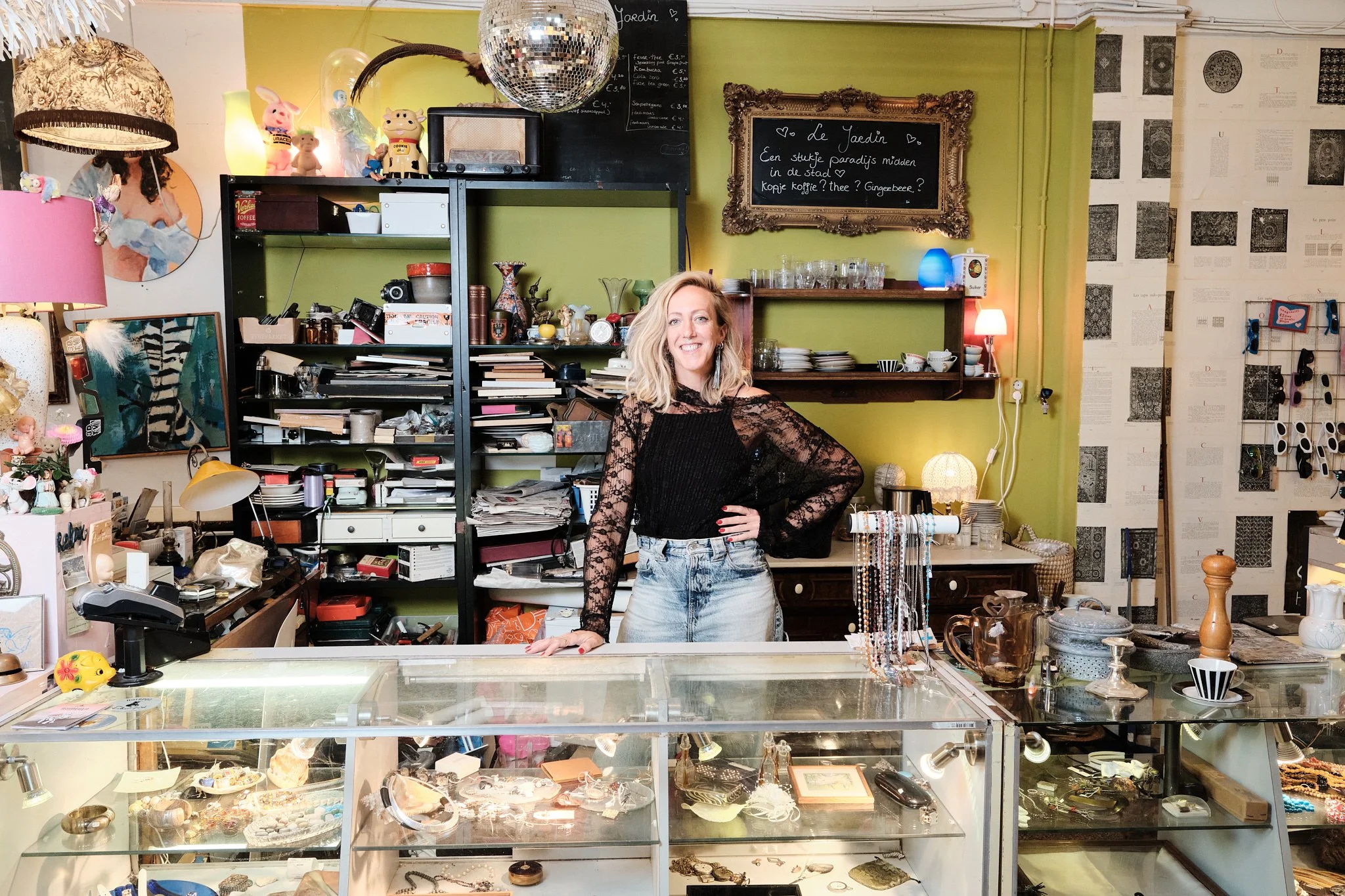 Een vrouw staat achter de toonbank van een antiek- en curiosawinkel met verschillende sieraden en accessoires. Op de achtergrond zijn schappen gevuld met boeken, decoratieve objecten en een zwarte poster met witgeschreven tekst.