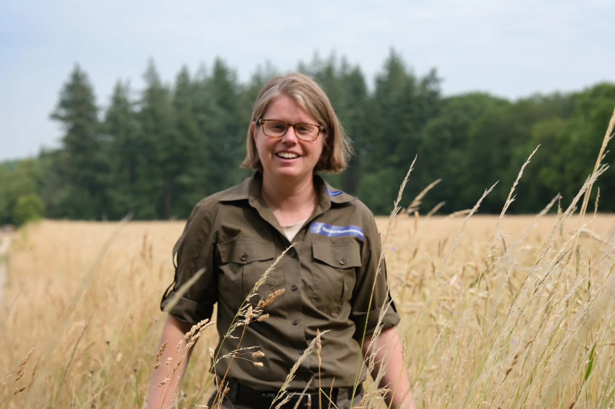 Vrouw in een boswachtersuniform in een akker met gras en bomen op de achtergrond.