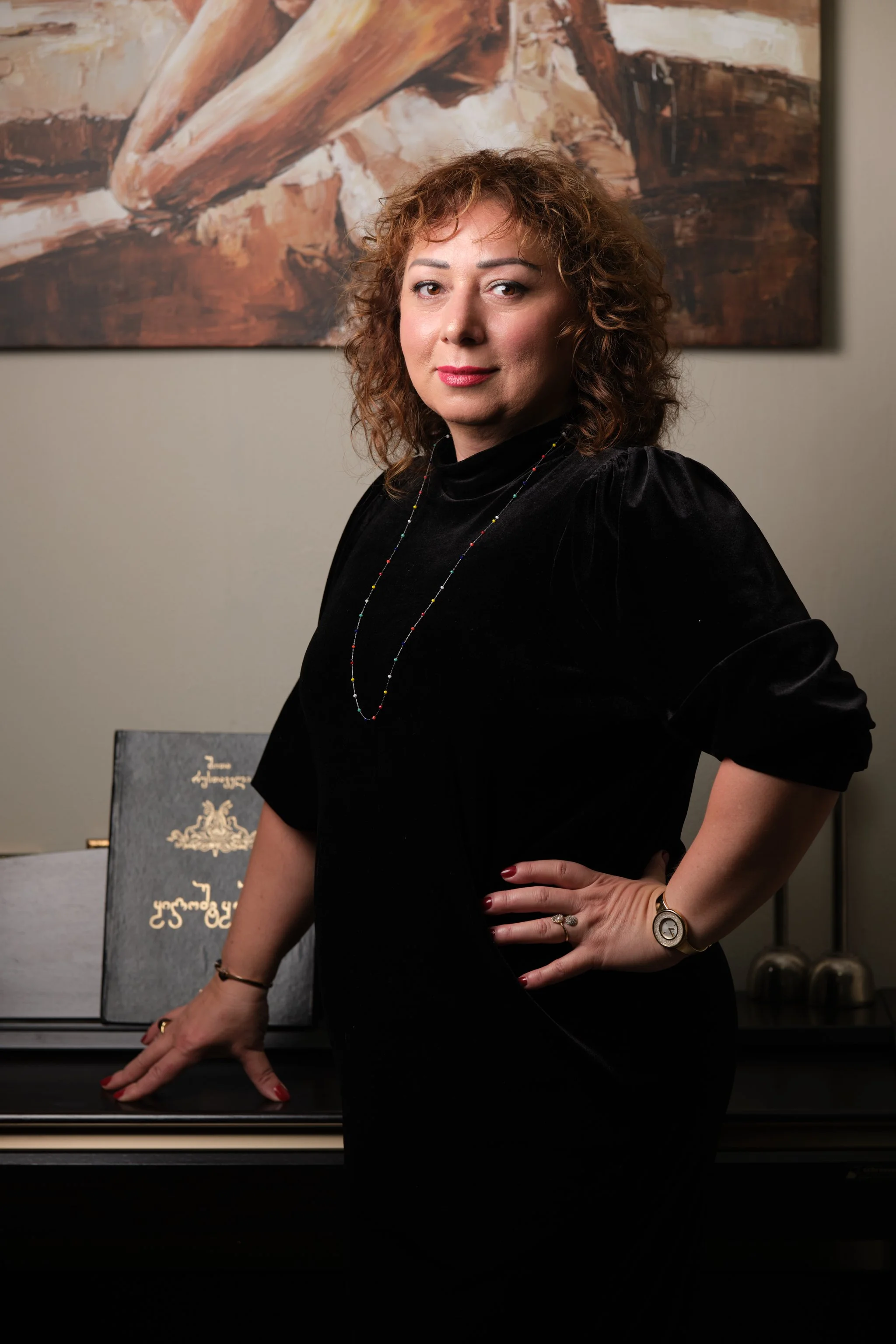 Een vrouw met bruin, krullend haar poseert voor een muur met een kunstwerk, naast een piano met een blad met Duitse tekst, gekleed in een zwarte jurk en sieraden. 