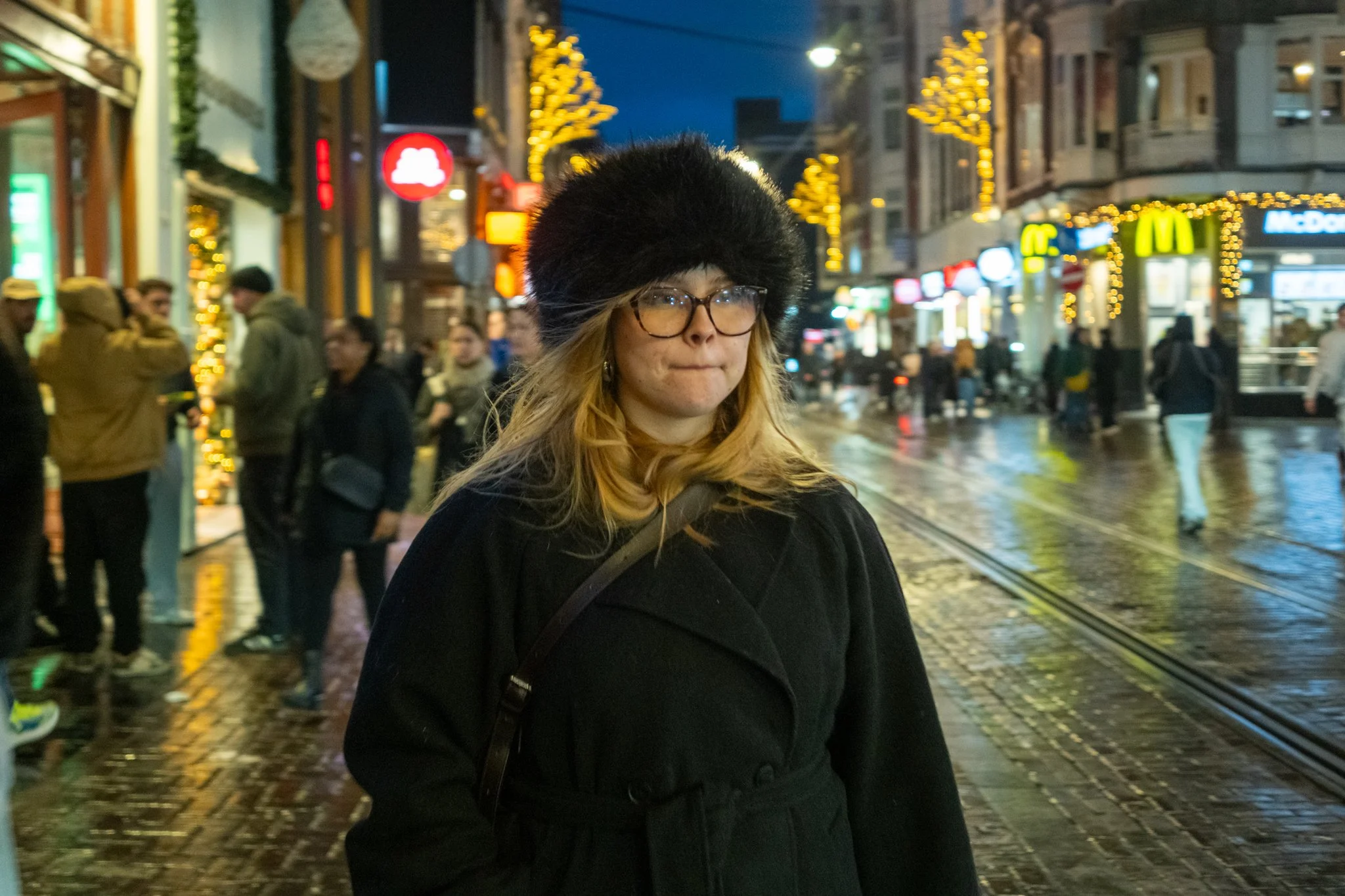 Een jonge vrouw met glazen en een bont muts staat op een stadsplein 's avonds, met kerstverlichting en winkels op de achtergrond.