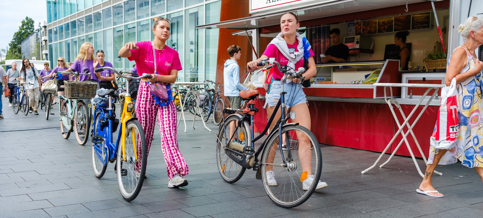 Vrouwen op fietsen in de stad, met een markt en mensen op de achtergrond.